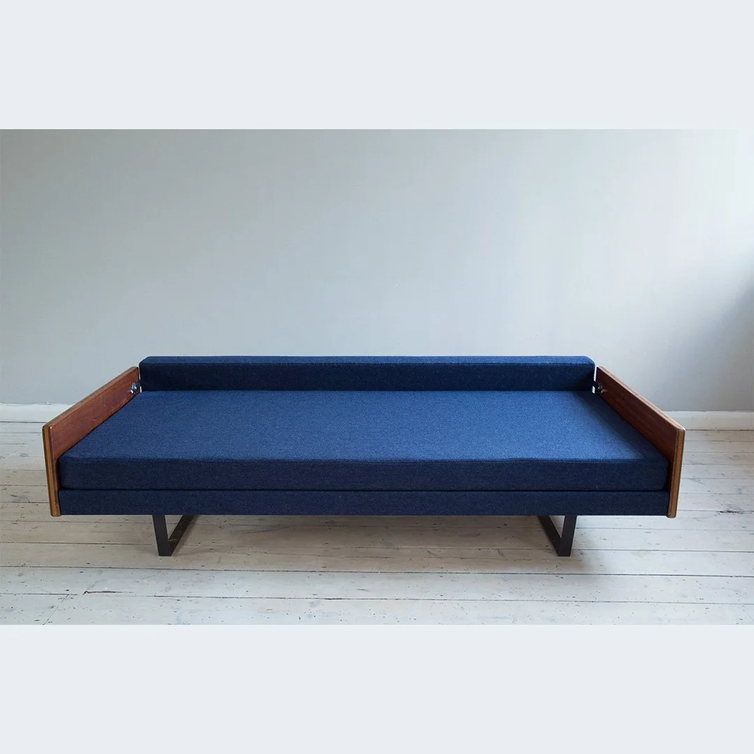 A mid-century modern sofa by Robin Day with dark blue wool upholstery fabric from Bute, wooden armrests, and black metal legs, placed against a light-coloured wall and white wooden floor.