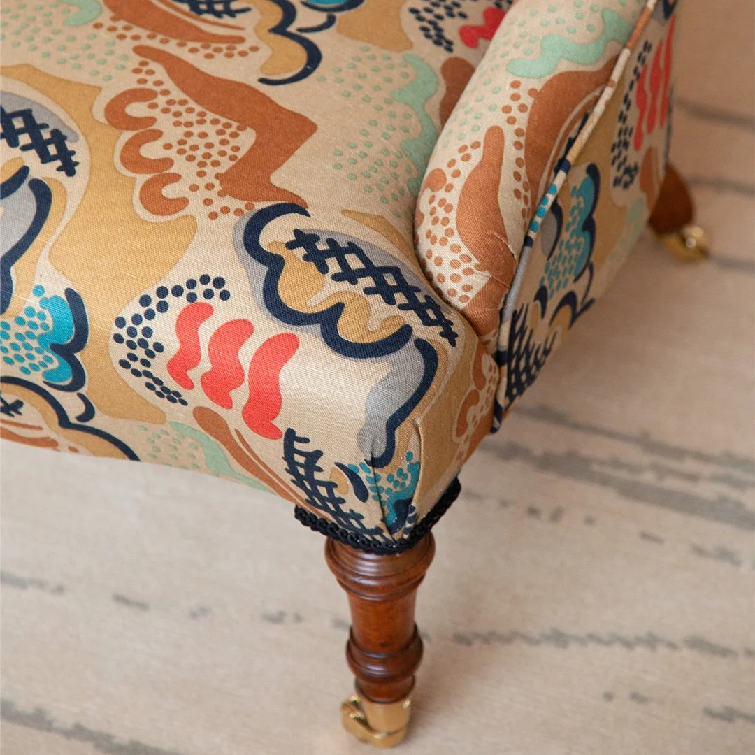 Close-up of a piece of furniture with a patterned fabric upholstery featuring abstract shapes in blue, red, beige, and black designed by Duncan Grant (Charleston), with a walnut leg with decorative carvings and brass castor wheels.
