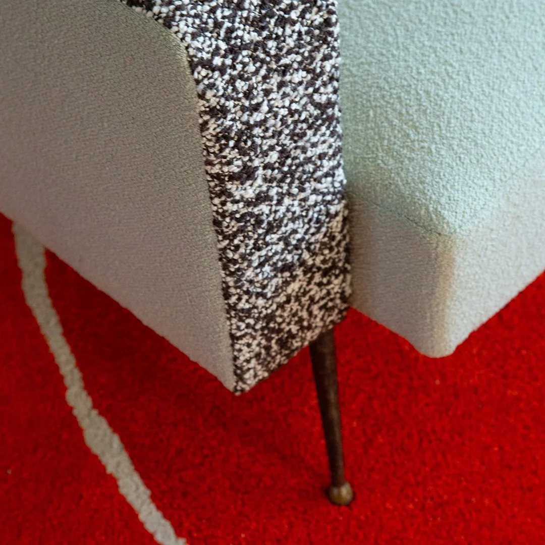Close-up of the corner of a light blue upholstered armchair with textured fabric, standing on a red carpet with white stripe.