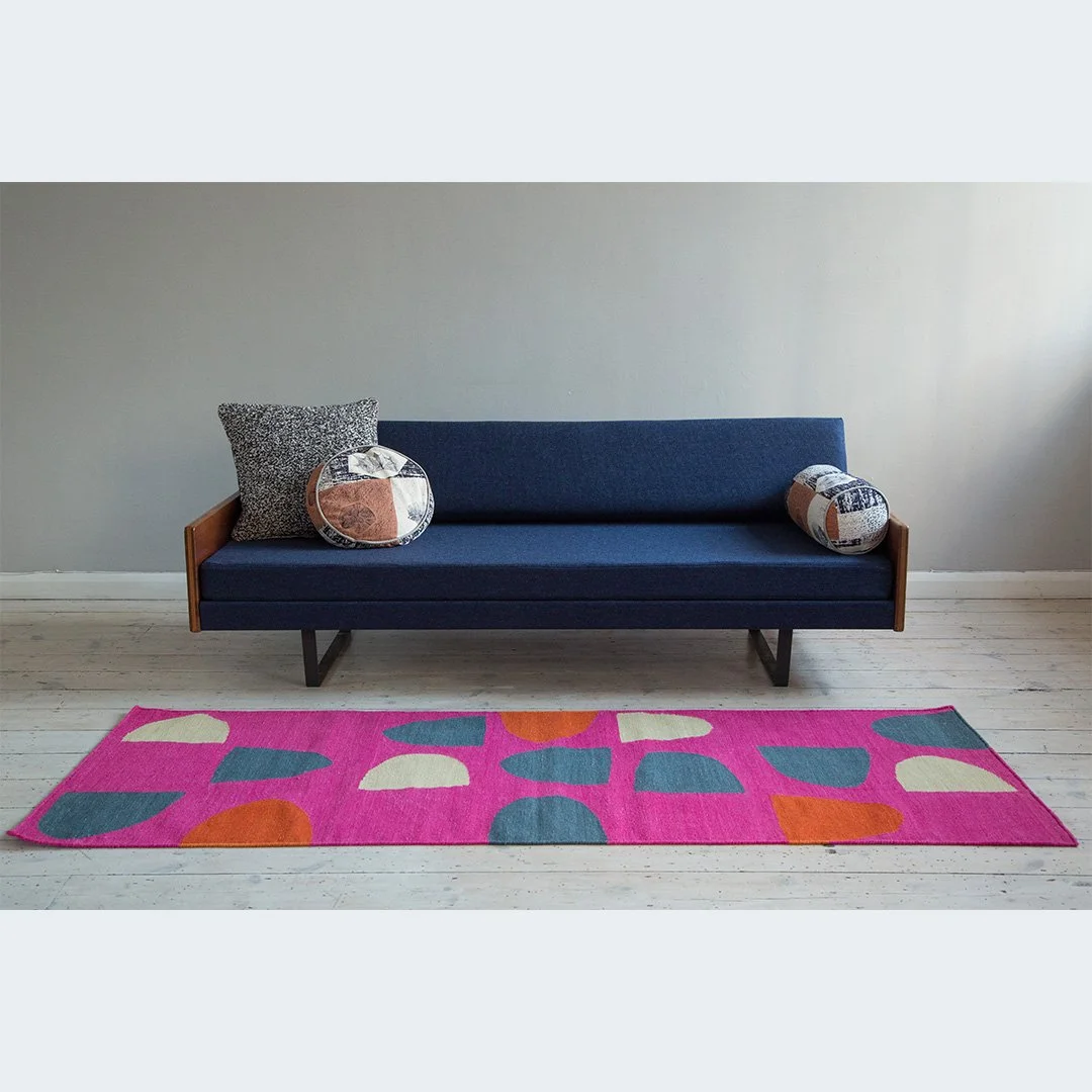 A minimalist living room with a navy blue Robin Day sofa, two patterned throw pillows, and a colourful pink rug with abstract shapes on light wood flooring, against a plain light-coloured wall.