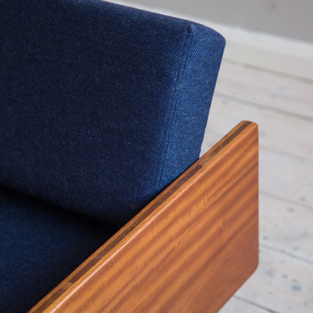 Close-up of a dark blue upholstered sofa / daybed designed by Robin Day with a wooden armrest, placed on a light-coloured wooden floor.