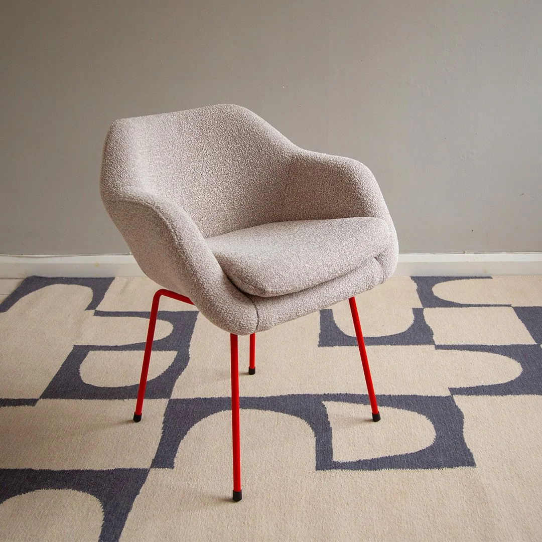 A Robin Day armchair upholstered in Bute wool boucle fabric with red metal legs sitting on a patterned beige and dark blue rug in front of a plain grey wall.