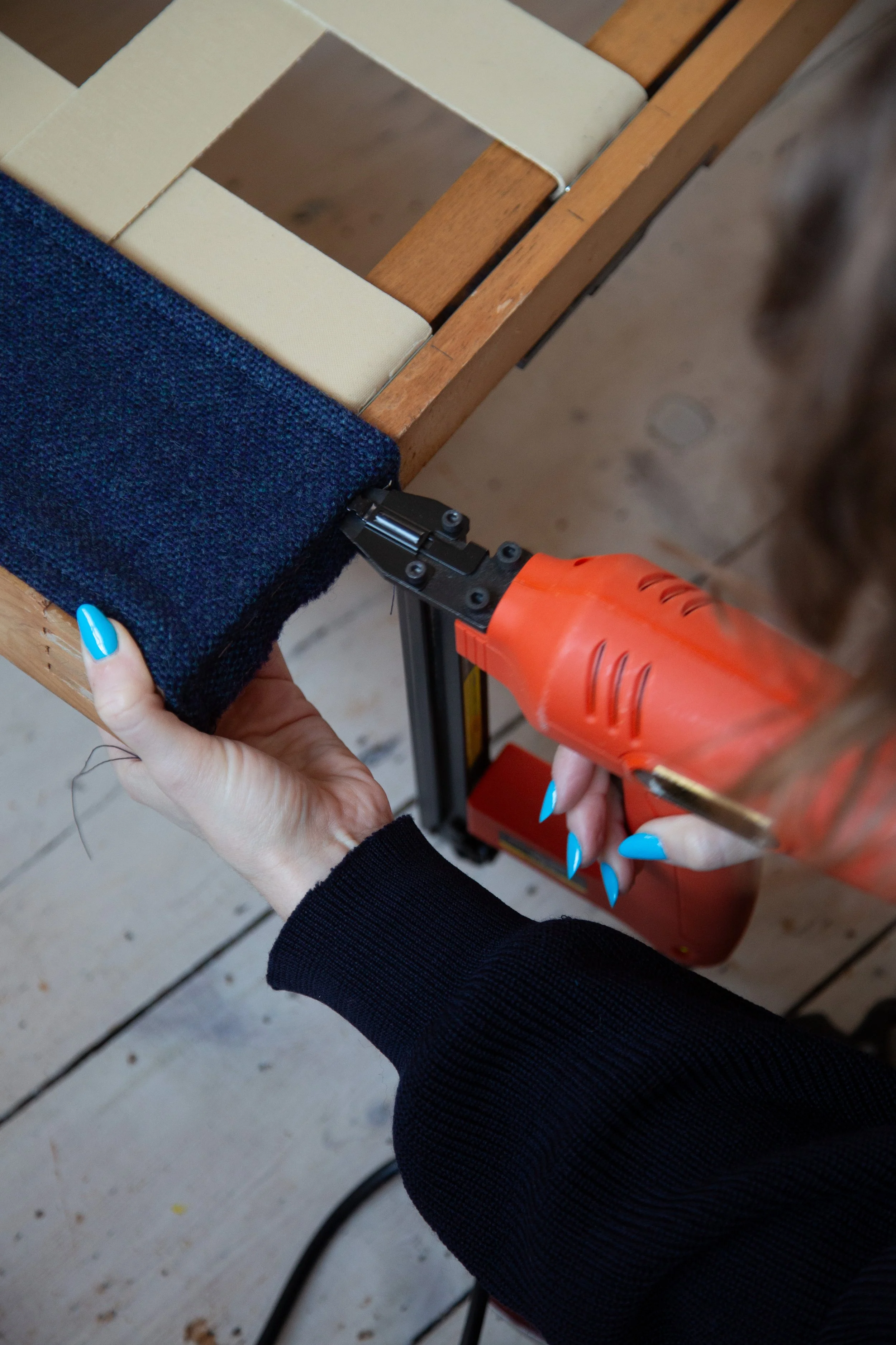Person using an orange staple gun to attach a piece of dark blue woollen fabric to a wooden sofa frame.
