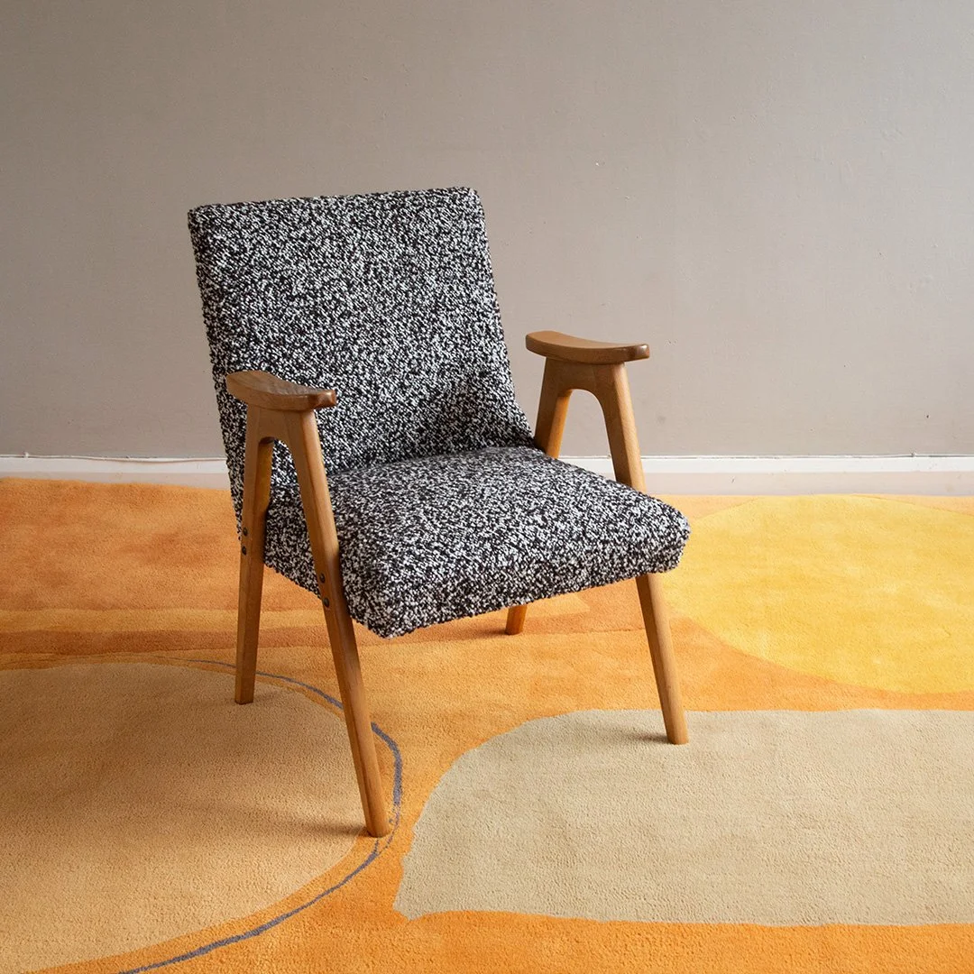 A Scandinavian mid-century modern armchair upholstered in wool boucle with wooden armrests and legs, placed on a colourful abstract-patterned rug in front of a plain wall.