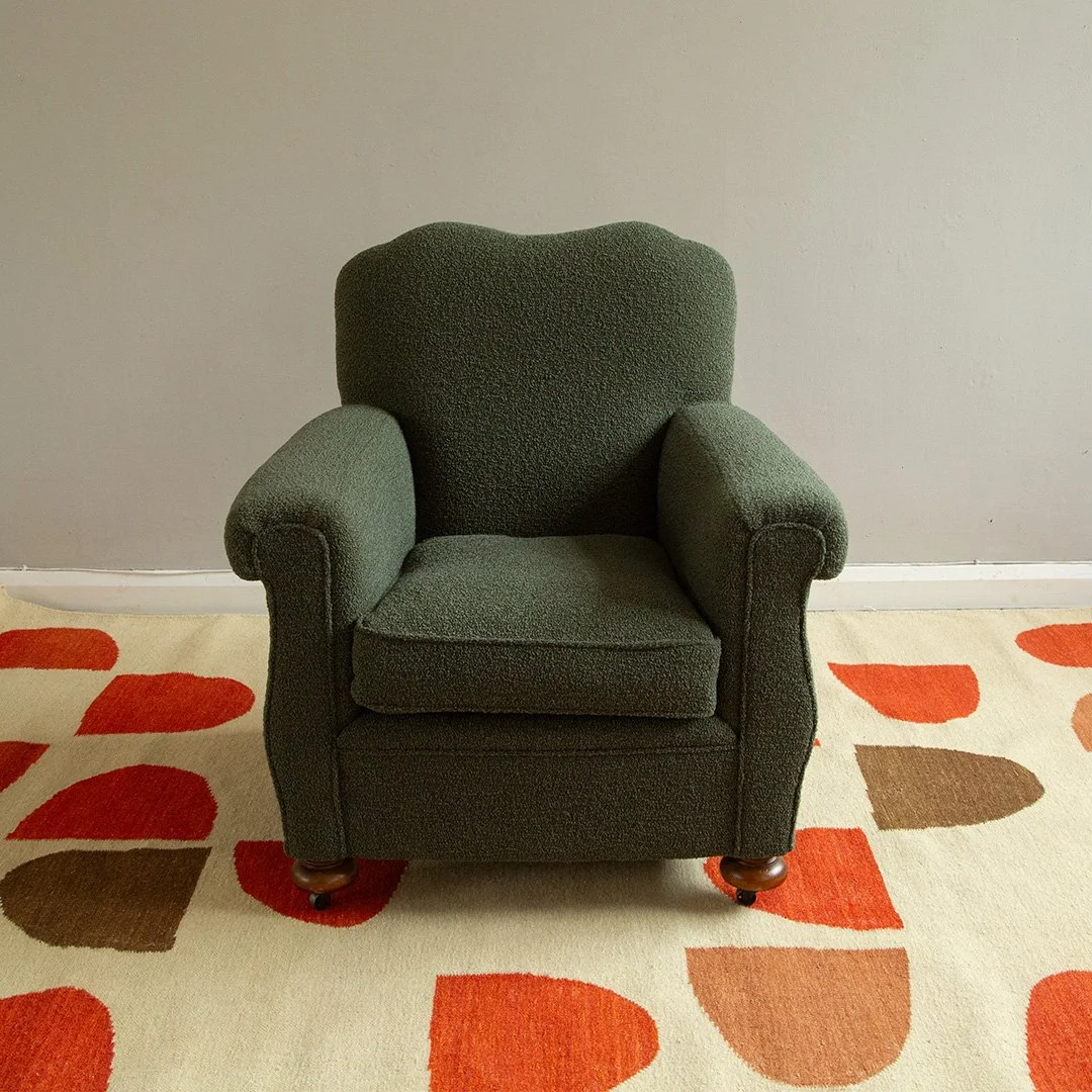A dark green armchair  upholstered in wool boucle from Designers Guild with rolled arms and wooden legs on a colorful rug with red, orange, and brown leaf-shaped patterns, against a plain gray wall.