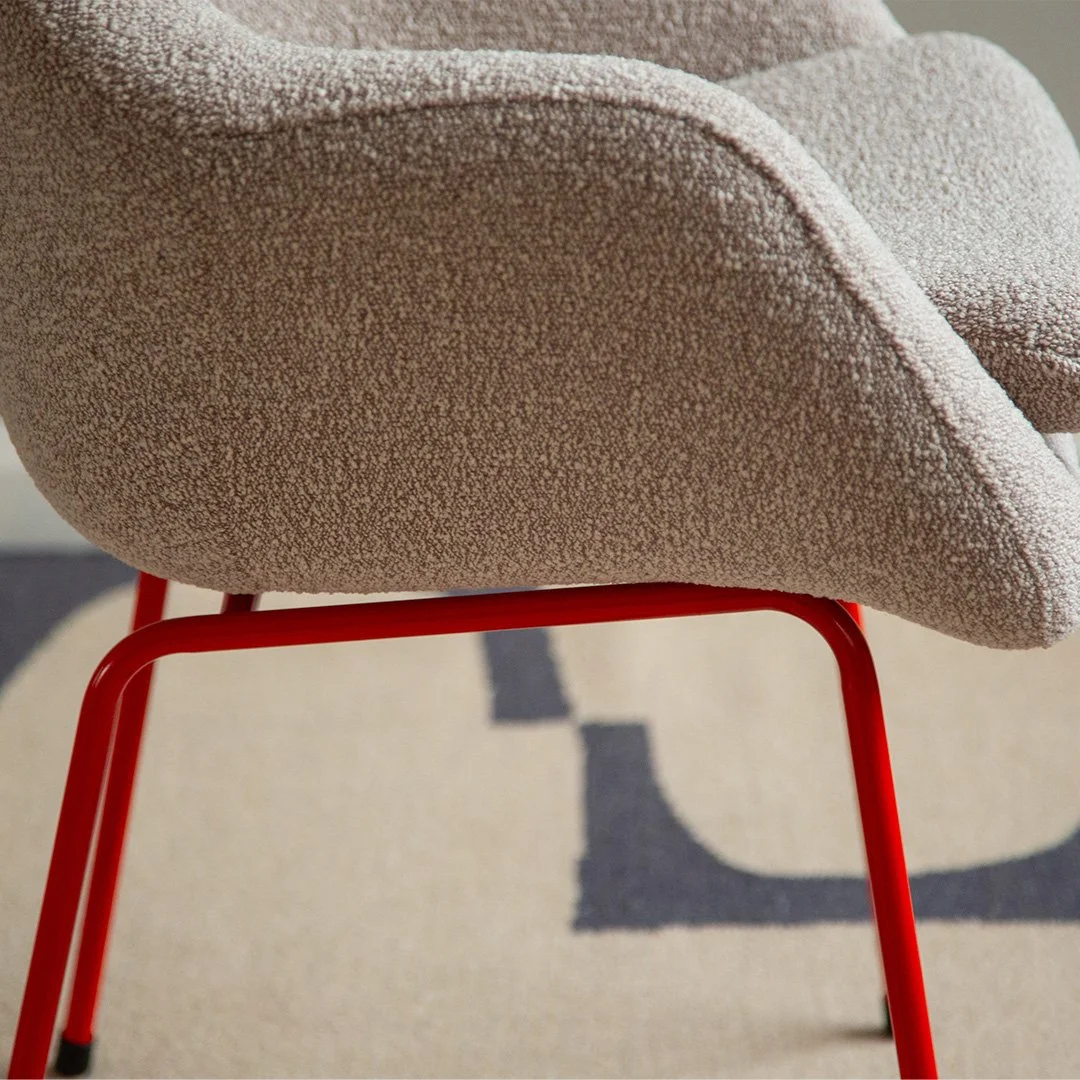 Close-up of a beige upholstered armchair with a vibrant red metal frame, situated on a patterned rug.