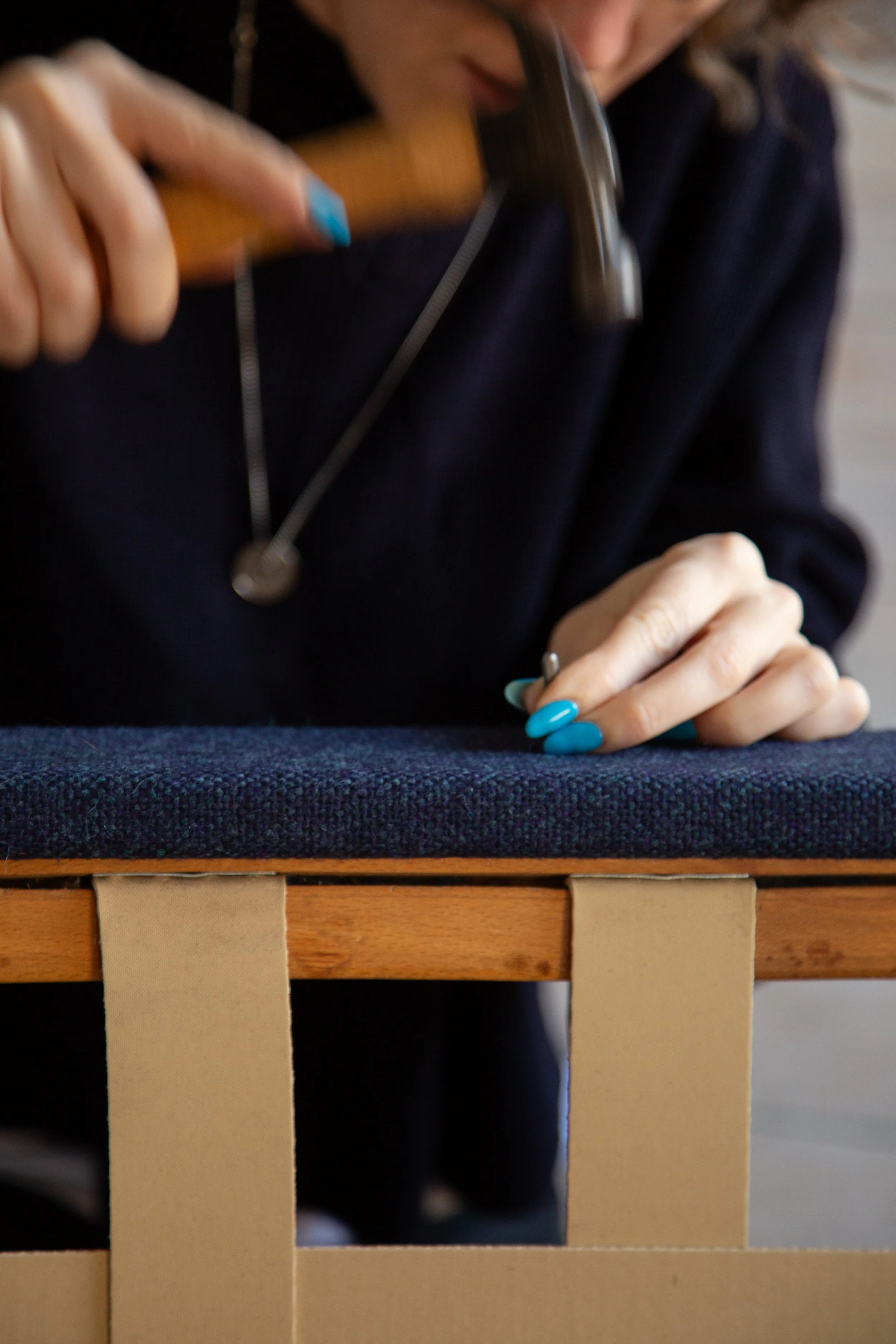 Close-up of an upholsterer person with blue nails hammering a tack into a piece of furniture, wearing a dark blue jumper.