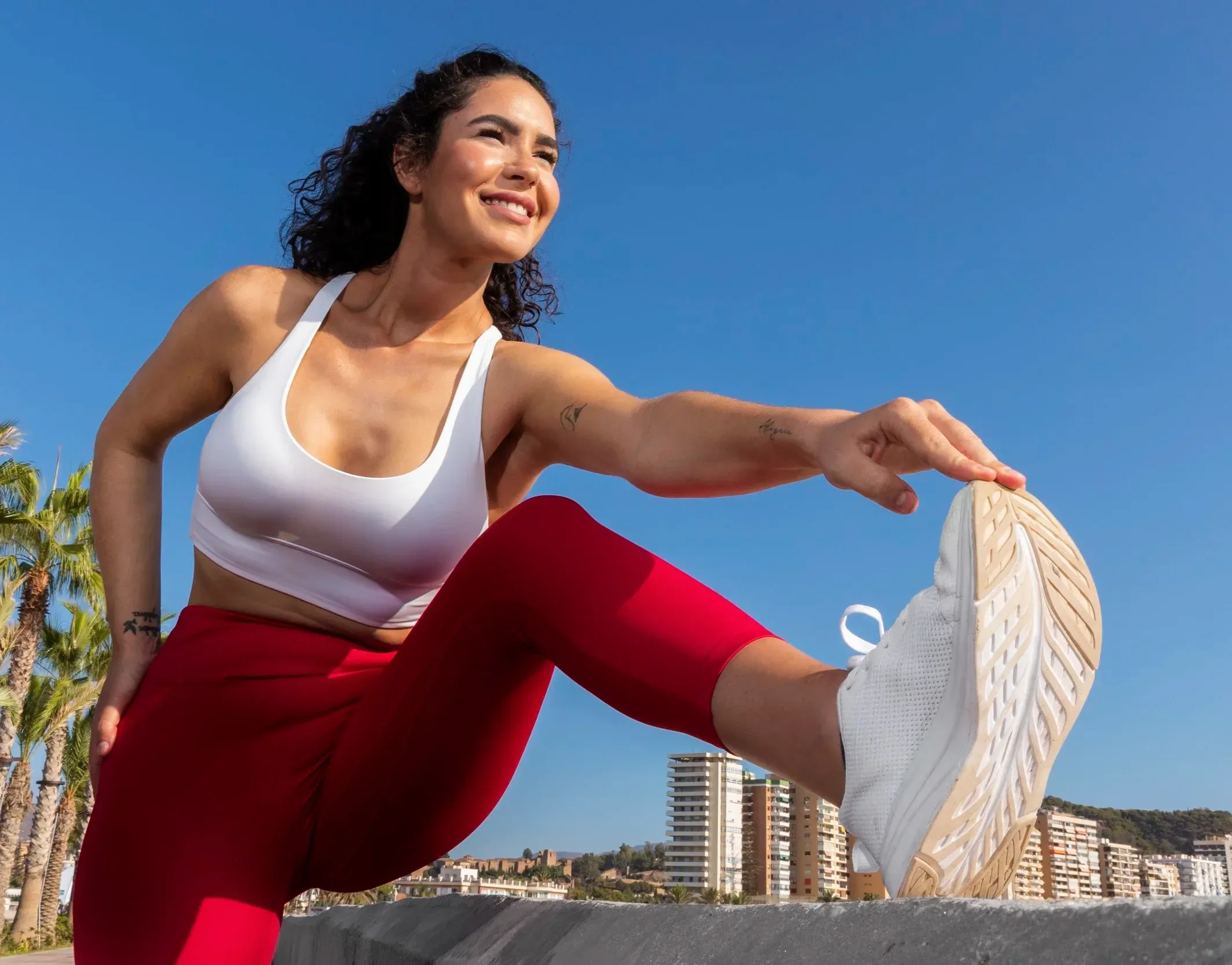 A woman in a white sports bra and red pants stretching outdoors with a cityscape and palm trees in the background. She is smiling and holding her foot up, showing her white athletic shoes.