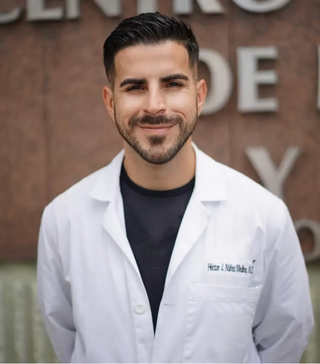 Dr. Héctor Núñez Medina smiling, wearing a white coat with the name 'Dr. Héctor Núñez Medina, MD' on it, standing outdoors in front of a brick wall.