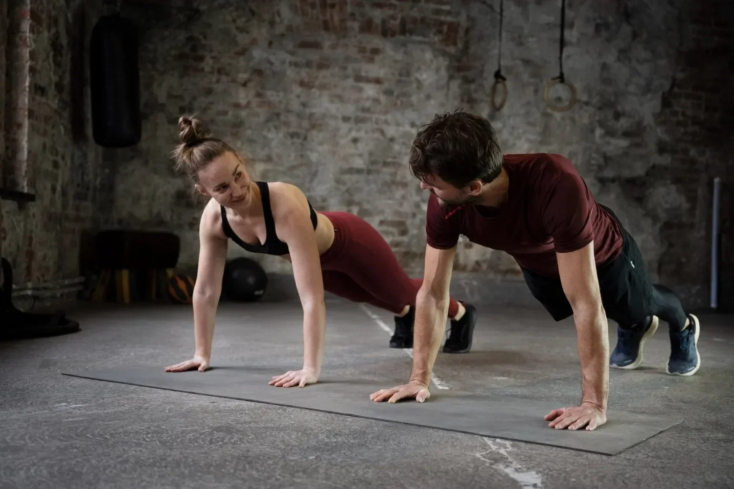 A woman and man are performing push-ups on a black exercise mat in a gym with a brick wall and fitness equipment in the background.