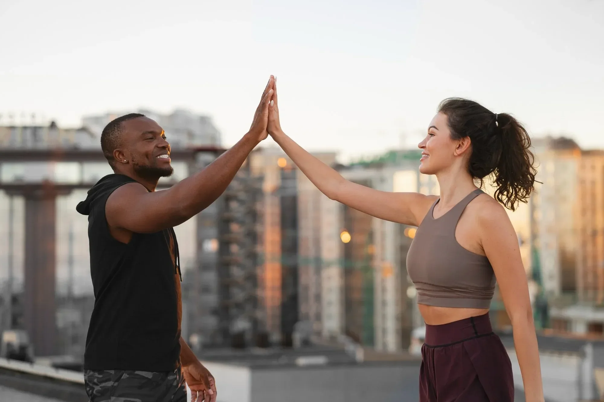 A man and woman are outdoors on a rooftop, giving each other a high five. The man is wearing a black hoodie and camouflage shorts, smiling. The woman is dressed in athletic wear, smiling back. In the background, there are buildings and a sunset sky.