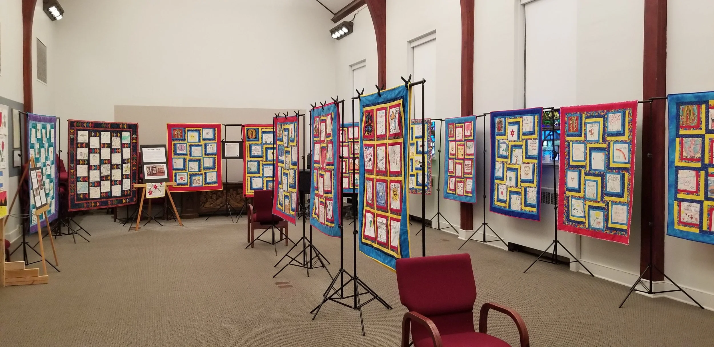 An indoor exhibit of colorful quilted wall hangings on black metal stands in a room with white walls and wooden beams.
