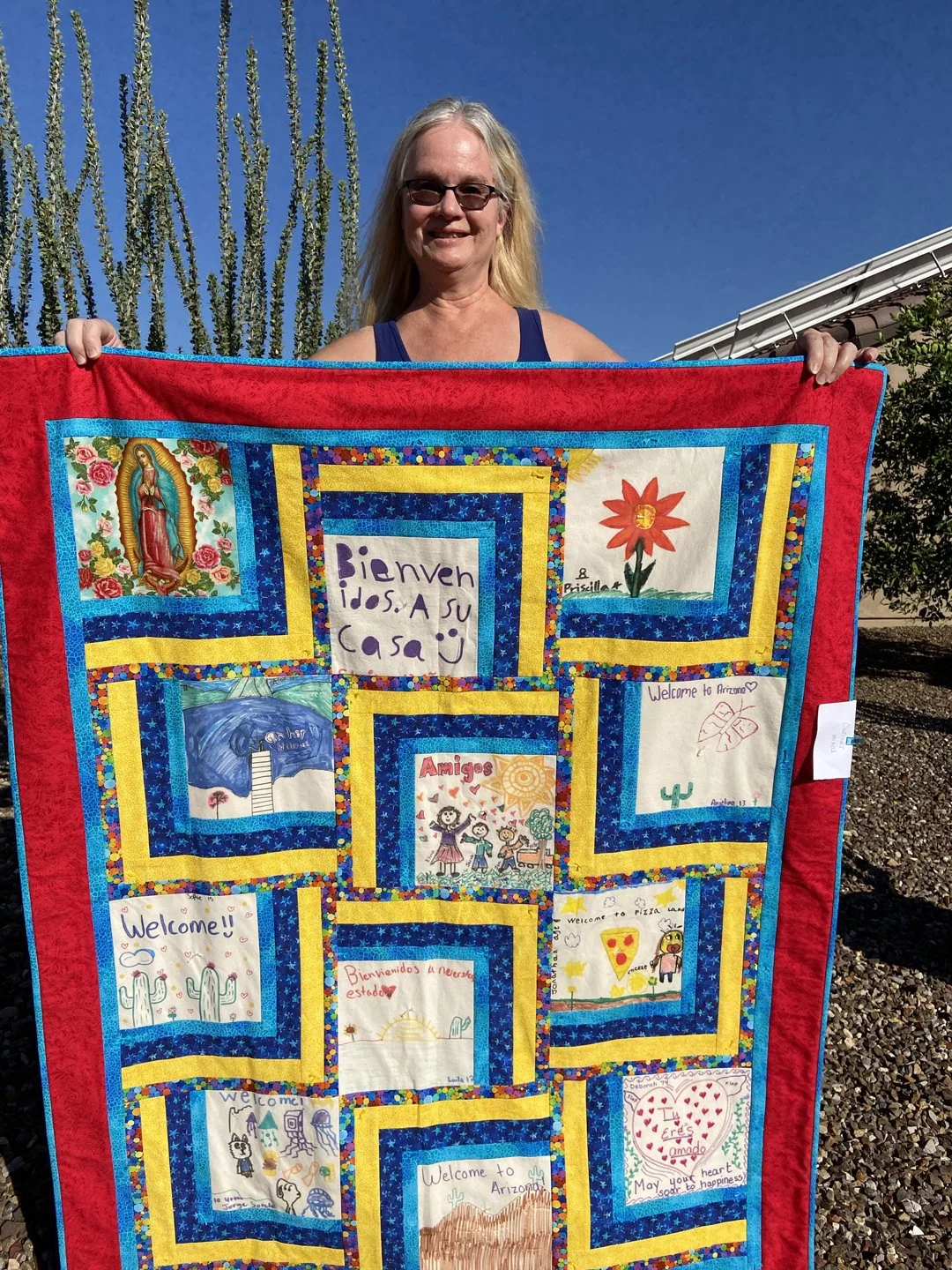 Gale Hall holding a colorful quilt with various drawings and messages, including welcome signs and artwork in Spanish and English.