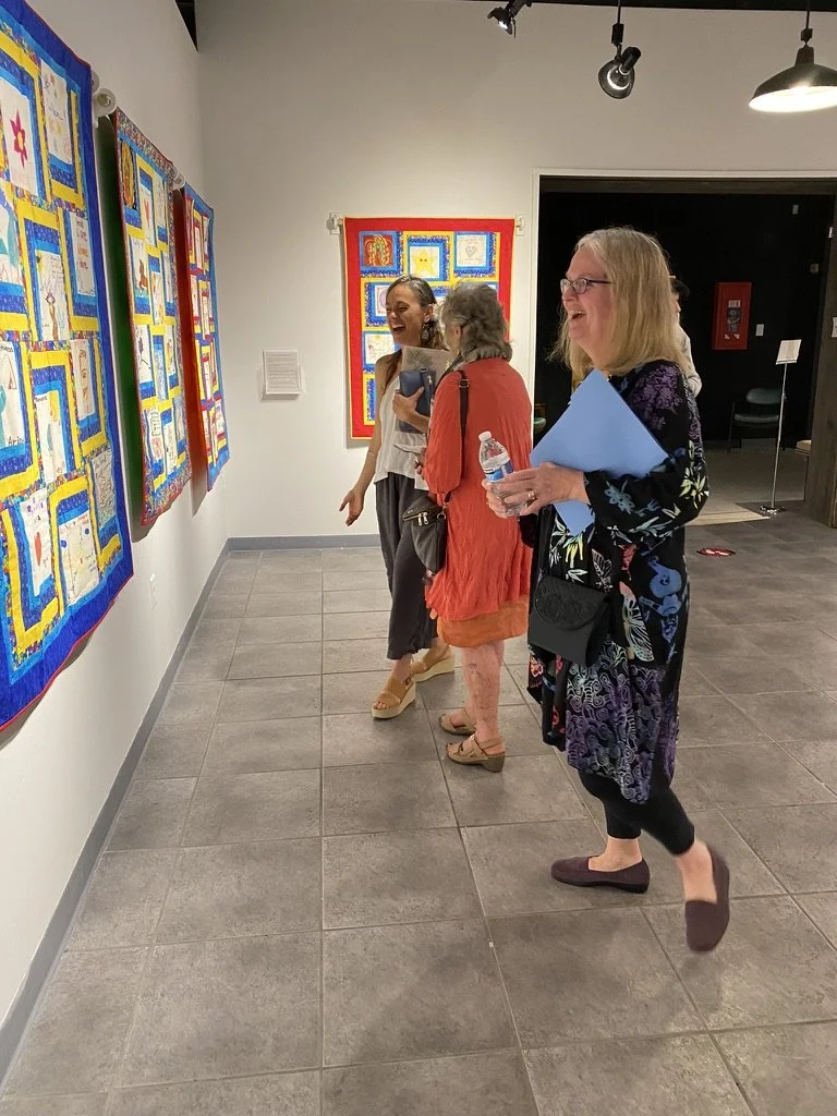 Three women examining colorful artwork in an art gallery, smiling and engaging with each other.