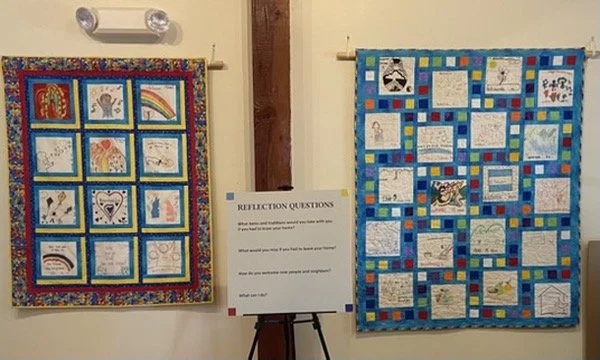 Two quilt panels with children's drawings and artwork, displayed on wall hooks, with a reflection sign in between them on an easel.