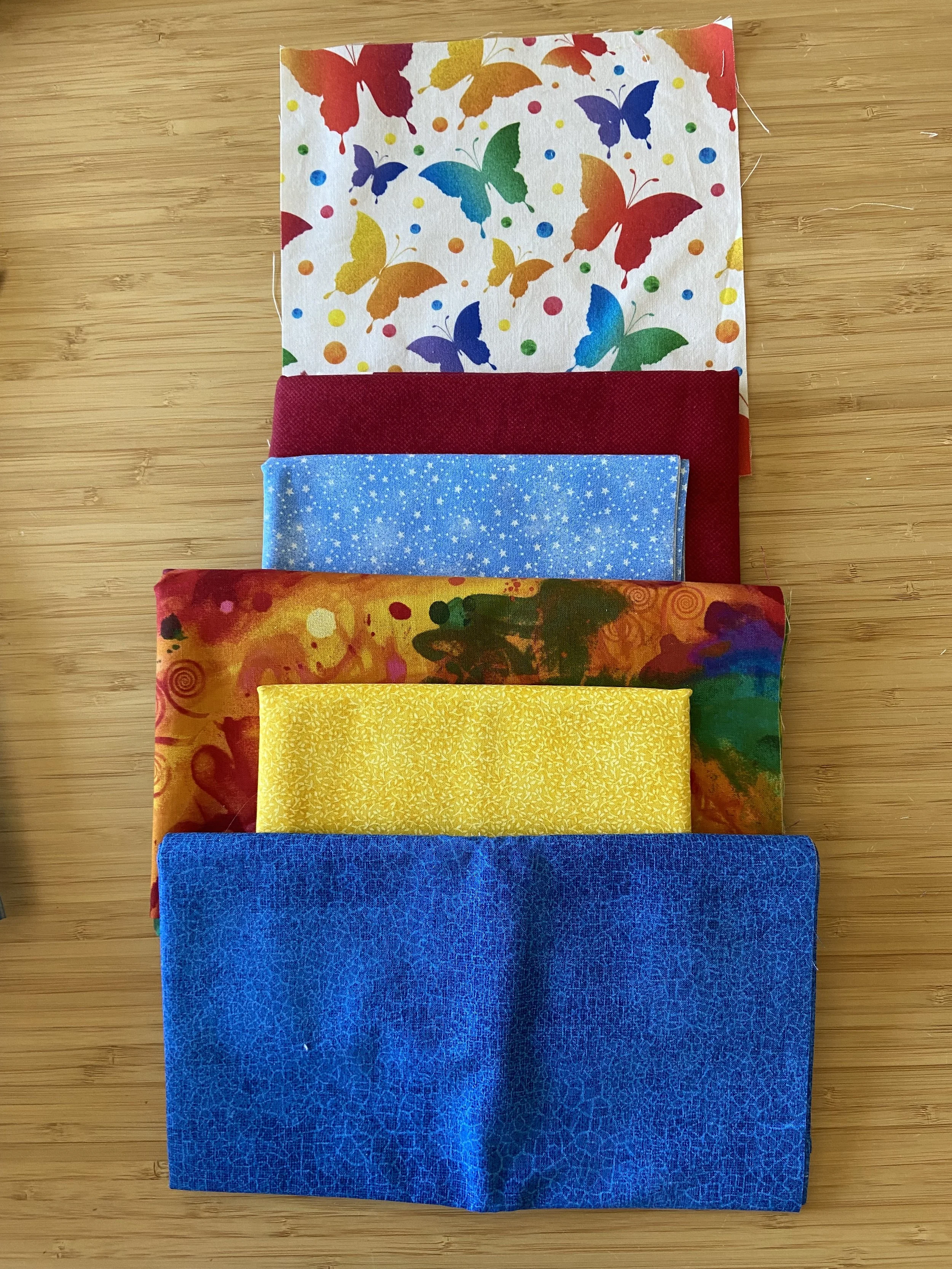 Six colorful single-use cloth face masks arranged vertically on a wooden surface, with patterns including butterflies, dots, and abstract designs.