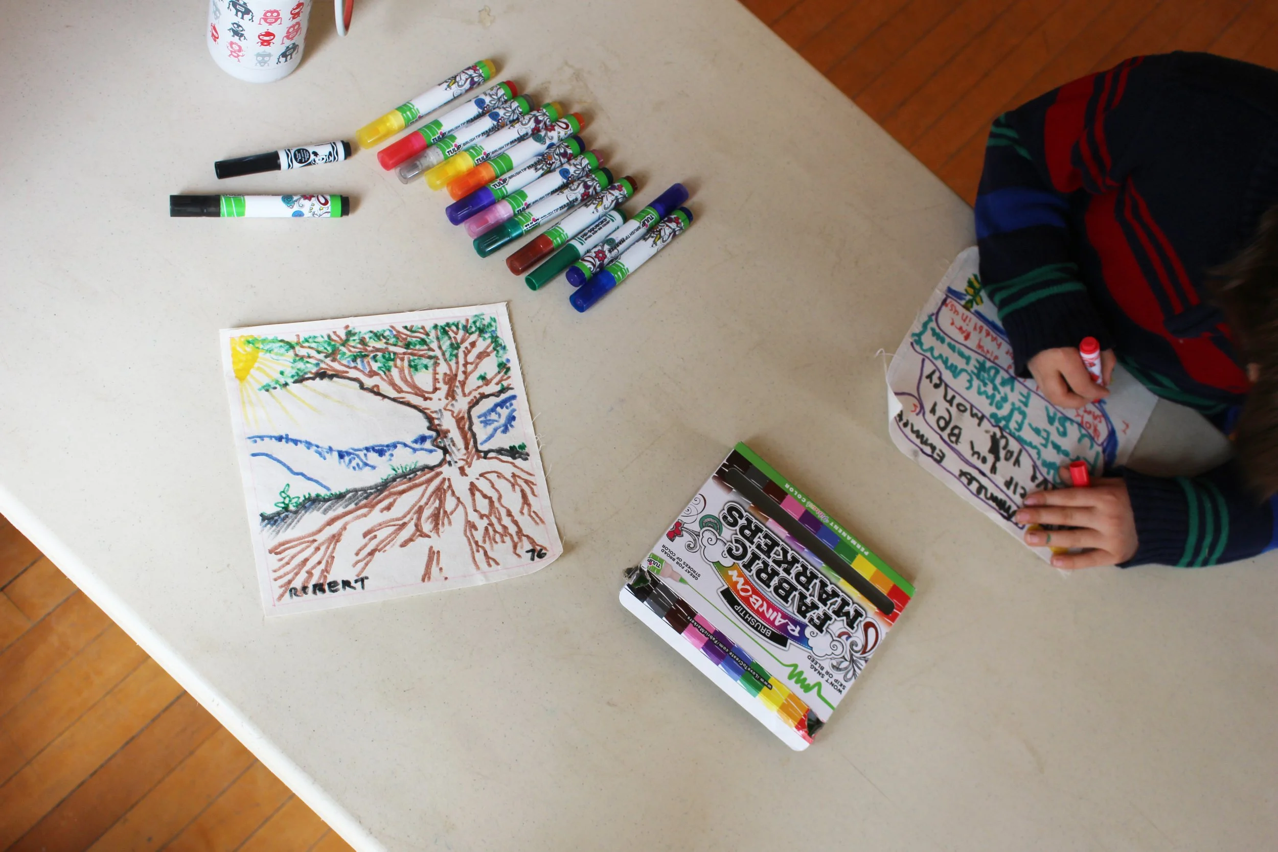 A table with a colorful drawing of a tree, a set of 12 colorful markers, and a partially visible person in a striped sweater writing on a paper or card.