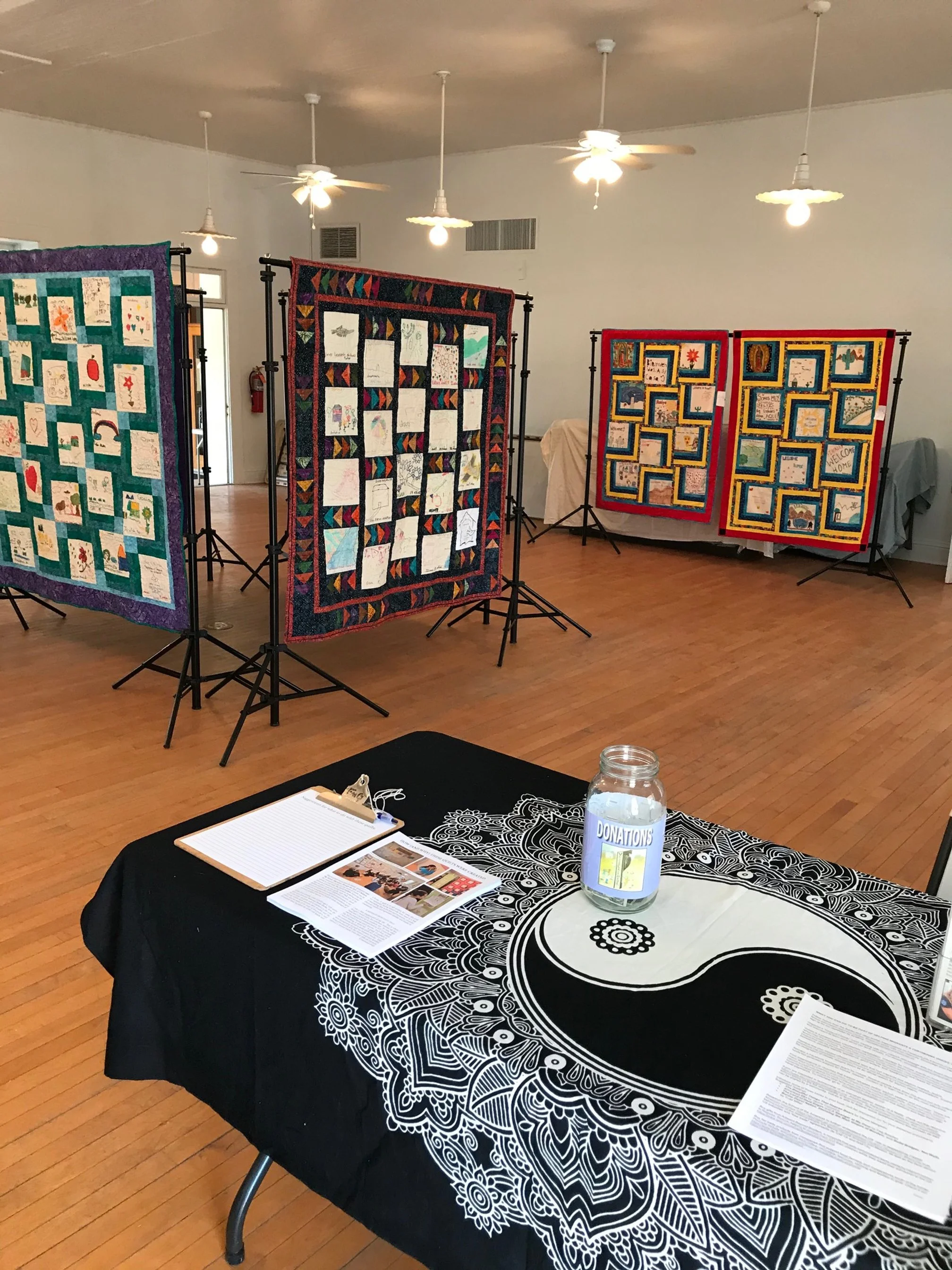 Indoor event with display panels showcasing artwork and writing, a table with a black cloth featuring a large white mandala design, a clipboard with papers, and a jar labeled 'Donations' on top of the table, with ceiling fans and bright lighting in the background.