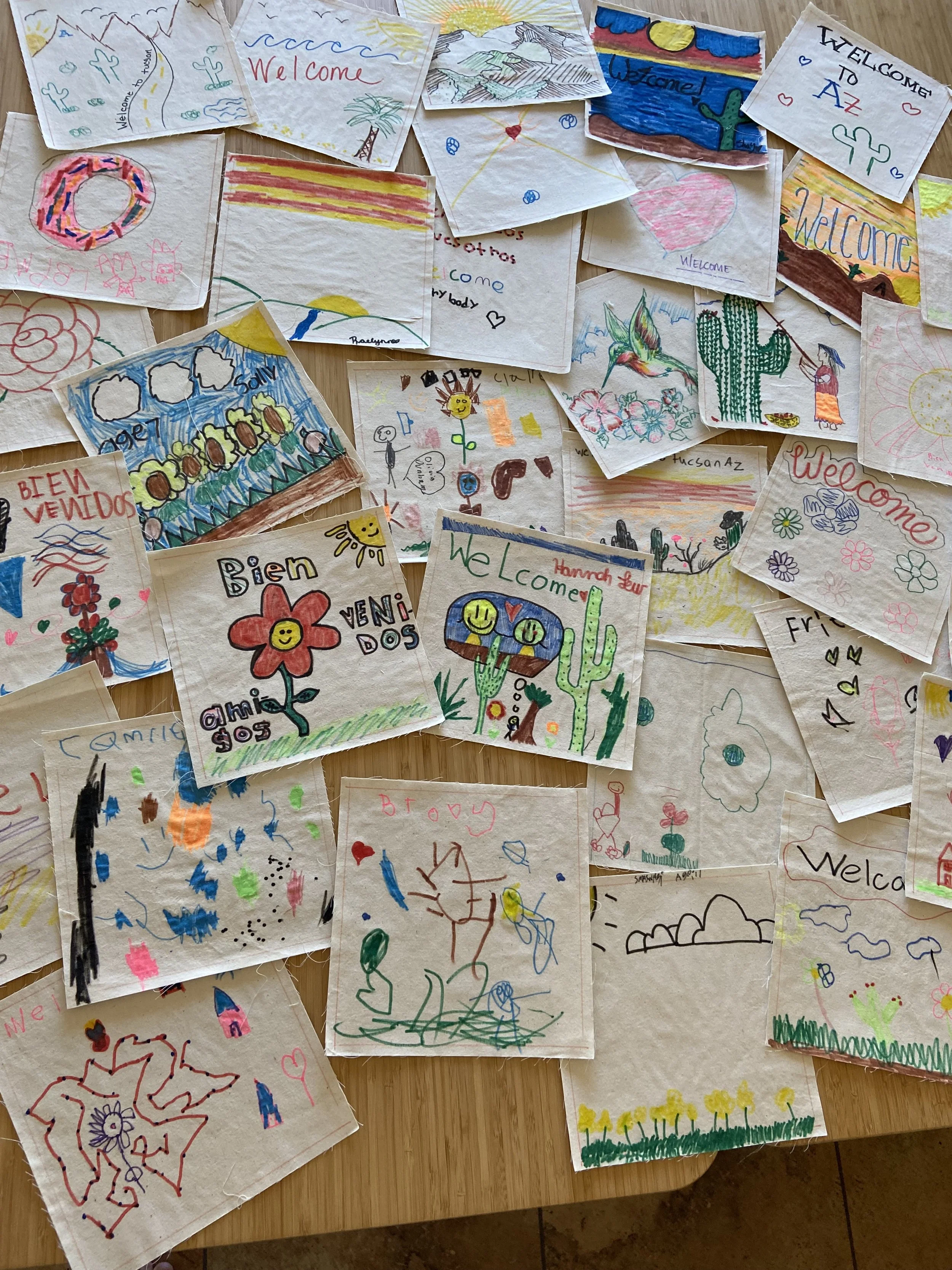 Several colorful children's drawings and handwritten notes on paper, featuring landscapes, welcome signs, flowers, hearts, animals, and various cheerful images, spread out on a wooden surface.