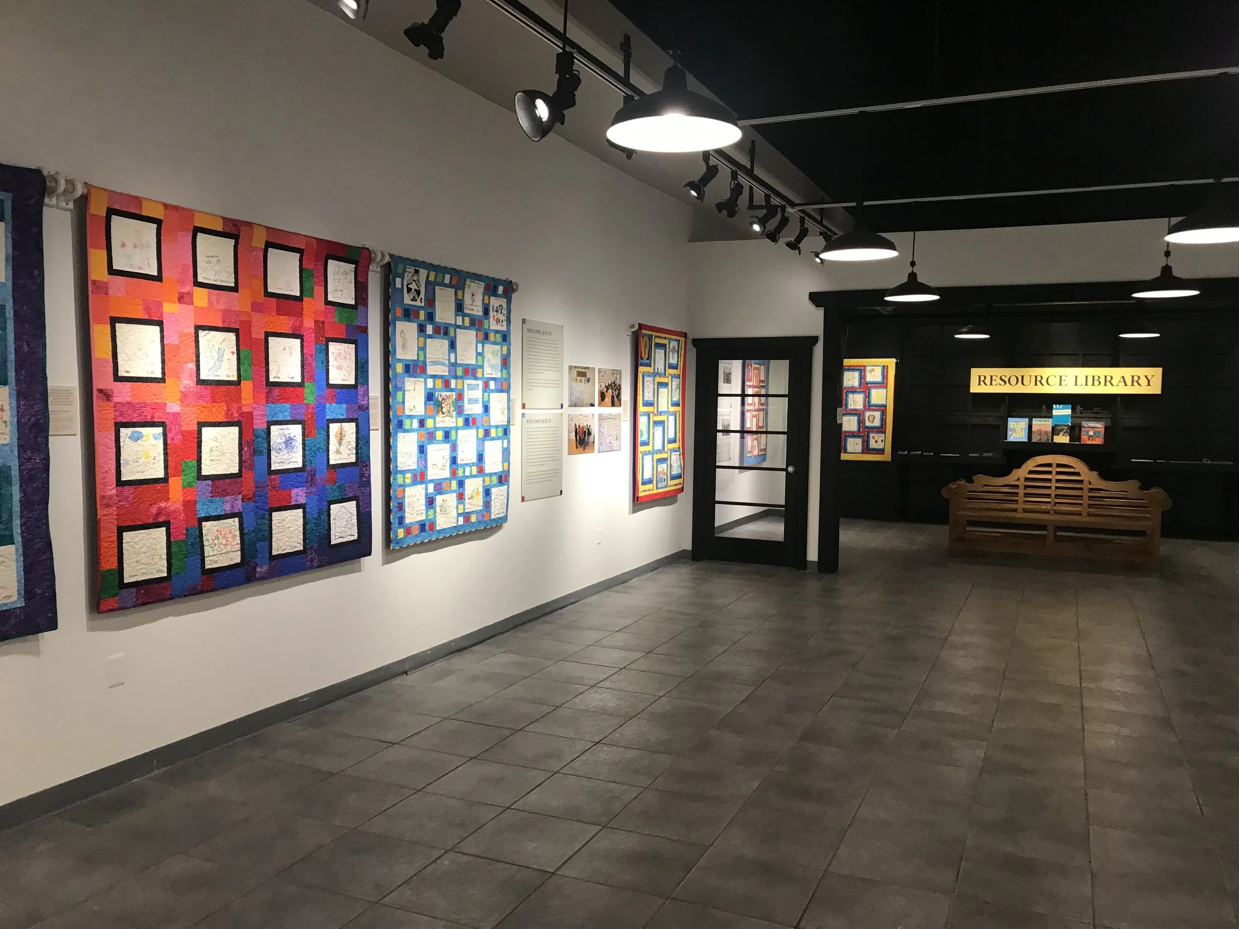 An indoor exhibition space displaying colorful quilts on the wall. There is a sign that reads 'Resource Library' near the door. The space is lit by overhead lights, and there is a wooden bench in the foreground.