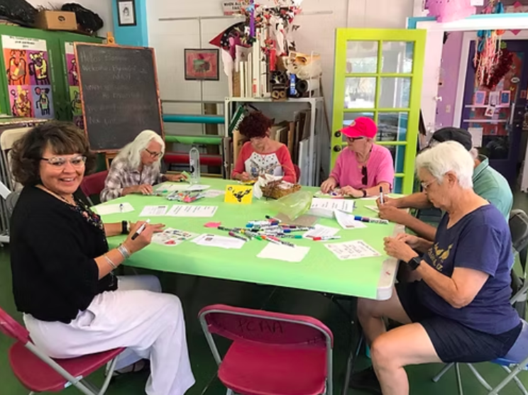 Group of women sitting around a bright green table engaging in arts and crafts activities in a colorful room.