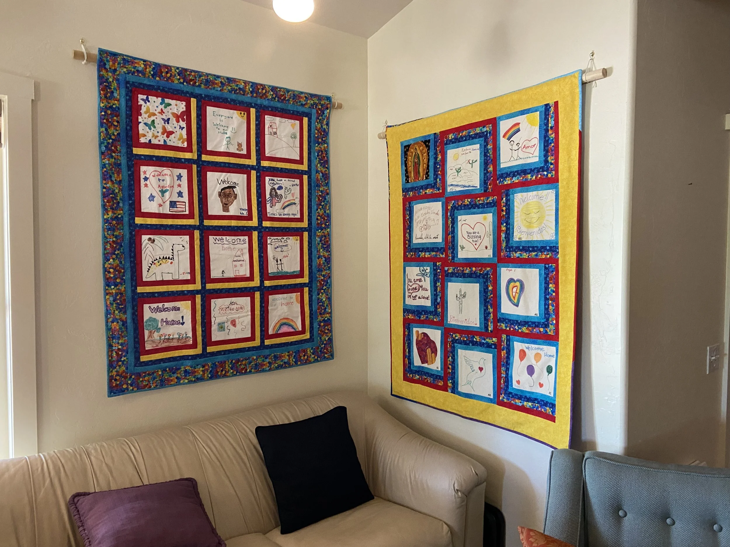 Two colorful quilts with children's drawings hang on the walls above a beige sofa with purple and black throw pillows in a living room.