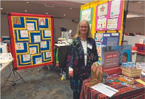 Gale Hall standing in front of colorful quilts at an indoor craft fair.