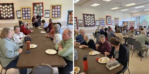 Group of elderly people sitting at tables in a community center or cafeteria, eating and conversing, with artwork displayed on the walls.