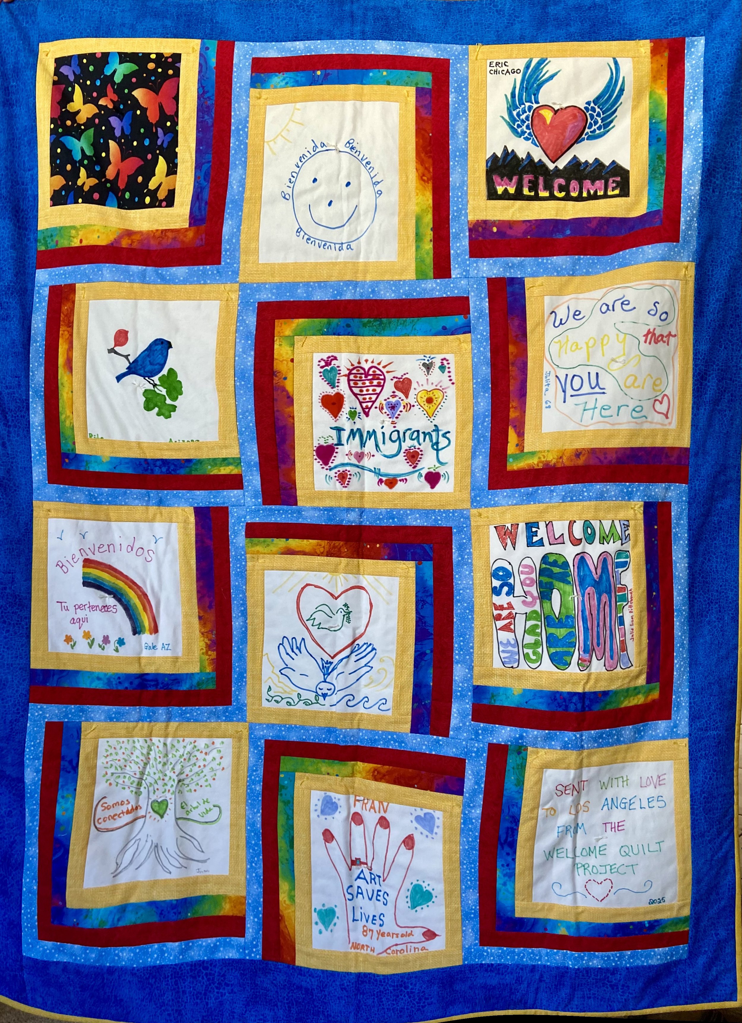 Colorful quilt with multiple embroidered and painted squares featuring messages such as "Welcome," "Immigrants," and "Art Saves Lives," along with illustrations of hearts, birds, rainbows, and other decorative designs, bordered with a blue starry fabric.