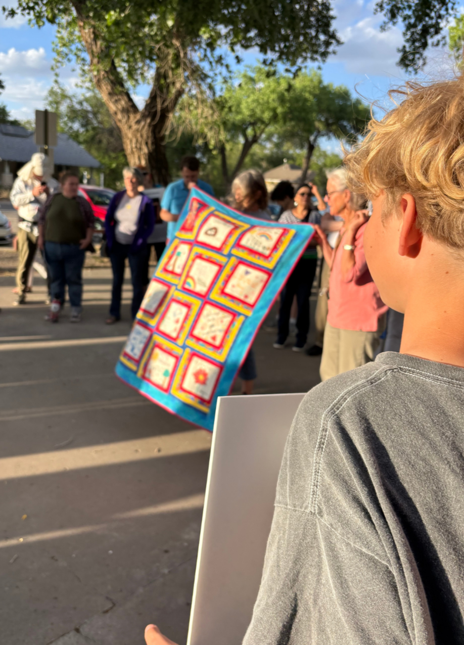 A group of people gathered outdoors, with a young boy in the foreground holding a blank sign or poster, while others hold a colorful quilt with various embroidered squares. The scene is set under trees with a sunny sky.