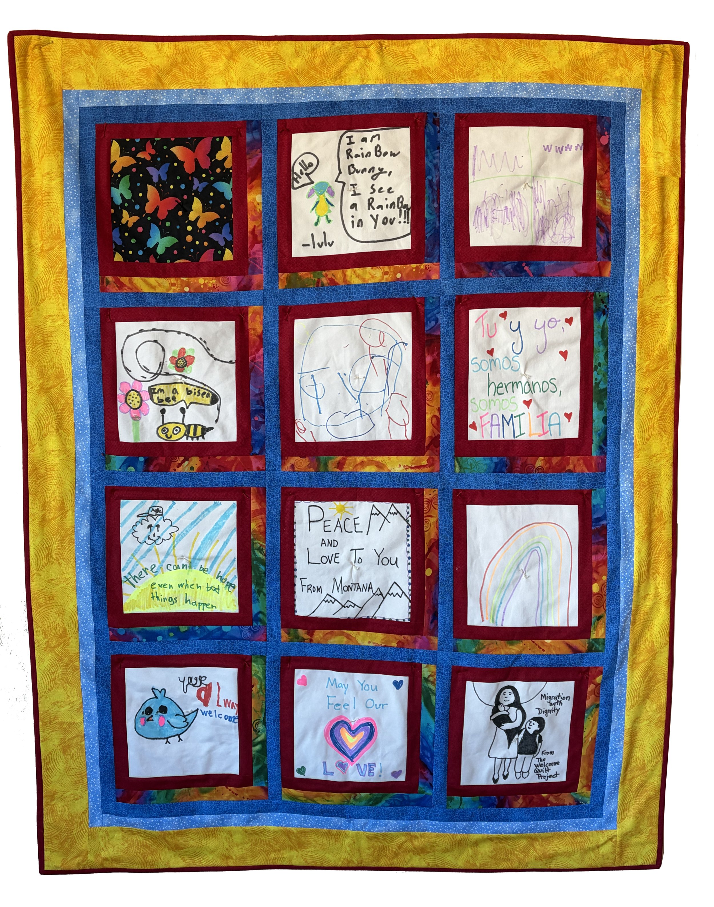 Colorful quilt with nine panels of children's drawings and messages, including rainbows, animals, and inspirational words, framed by yellow, blue, and red borders.
