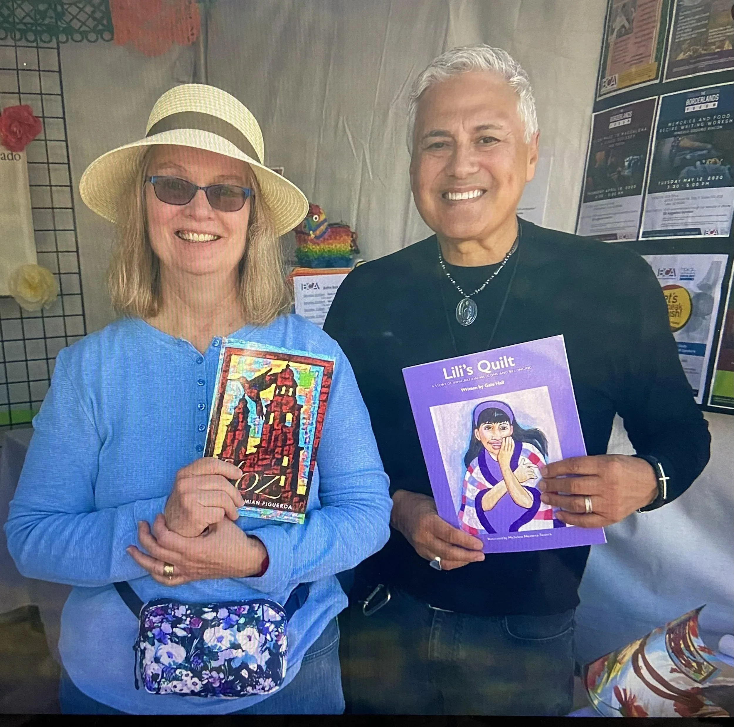 A woman wearing a yellow hat and glasses holding a colorful quilt book, and a man with gray hair holding a children's book titled "Lili's Quilt" at an indoor event.