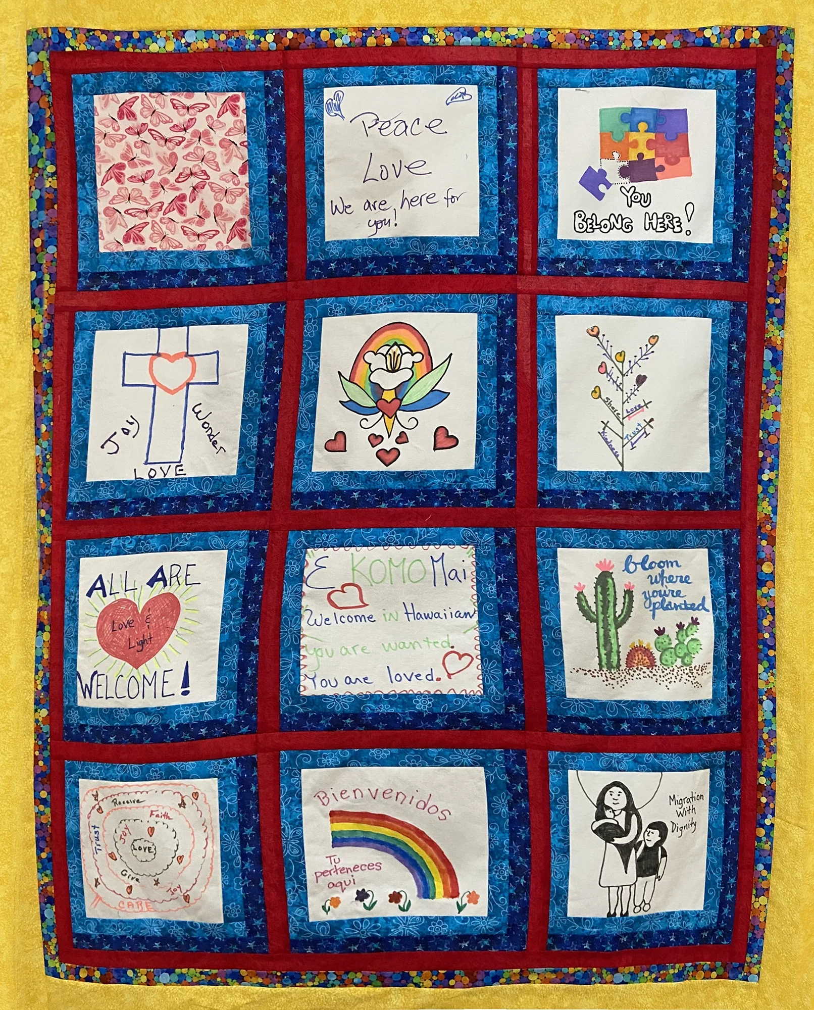 A colorful quilt display with various handmade posters and artwork inside a red-bordered frame. The posters feature messages of welcome, love, peace, equality, and diversity, with drawings of hearts, rainbows, a cross, a cactus, and people, set against bright and cheerful backgrounds.