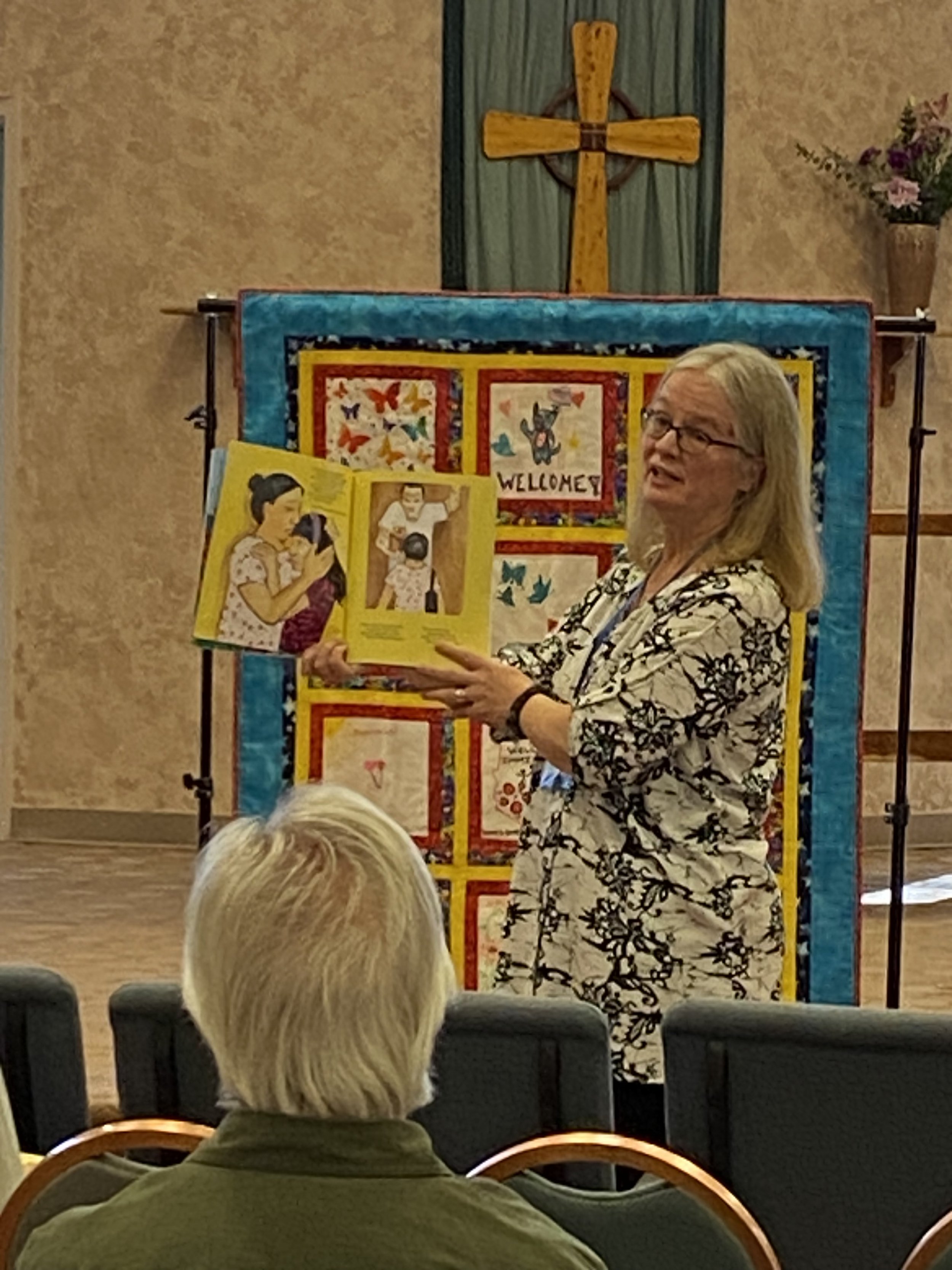 Gale Hall holding an open book with colorful illustrations and text that says 'Welcome' is standing in front of a quilt and a cross on a green and beige wall, speaking to an audience.