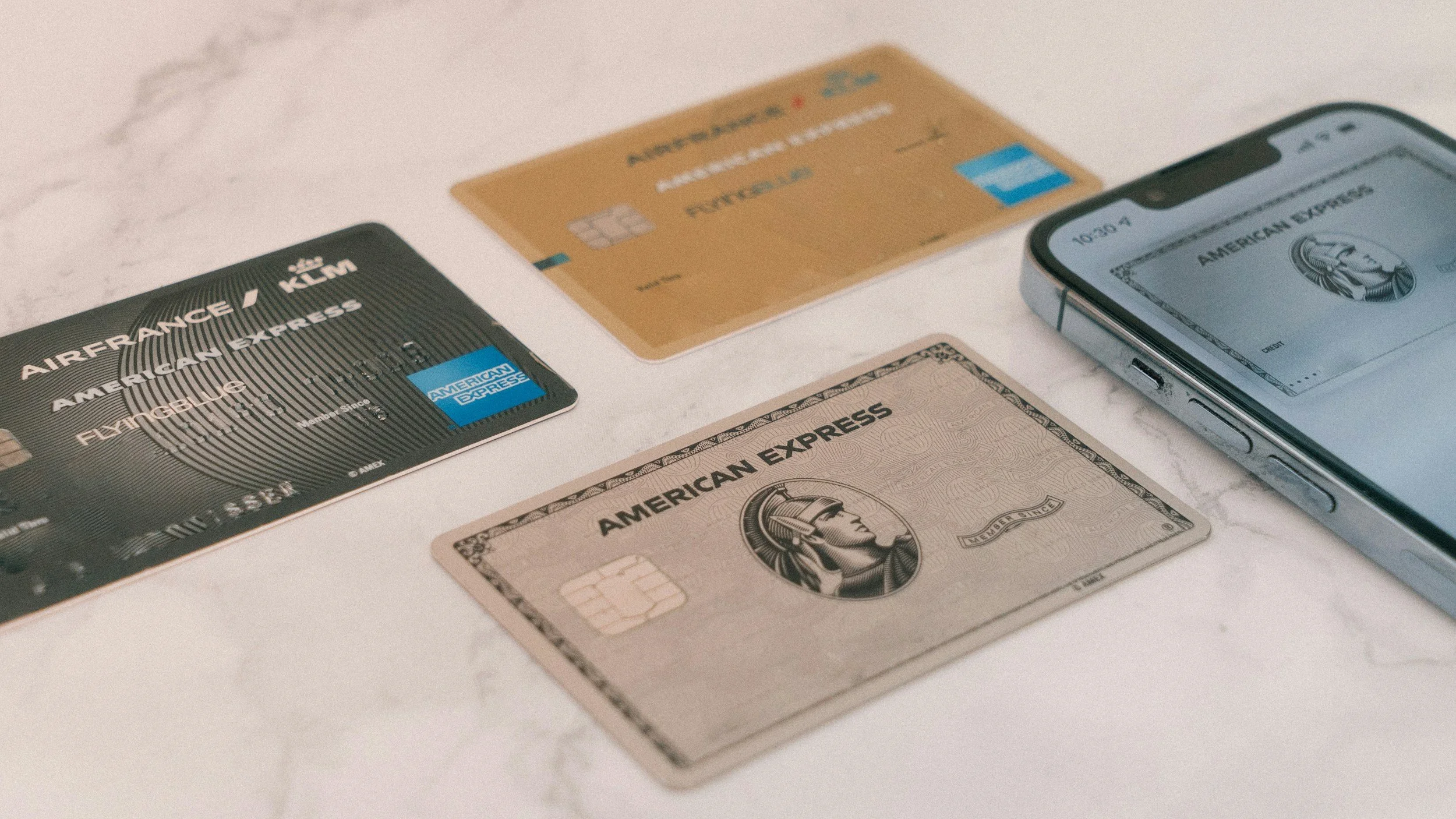 An American Express black credit card placed on a white marble surface, with a gold credit card, an American Express white card, a fifty-dollar bill, and a smartphone nearby.