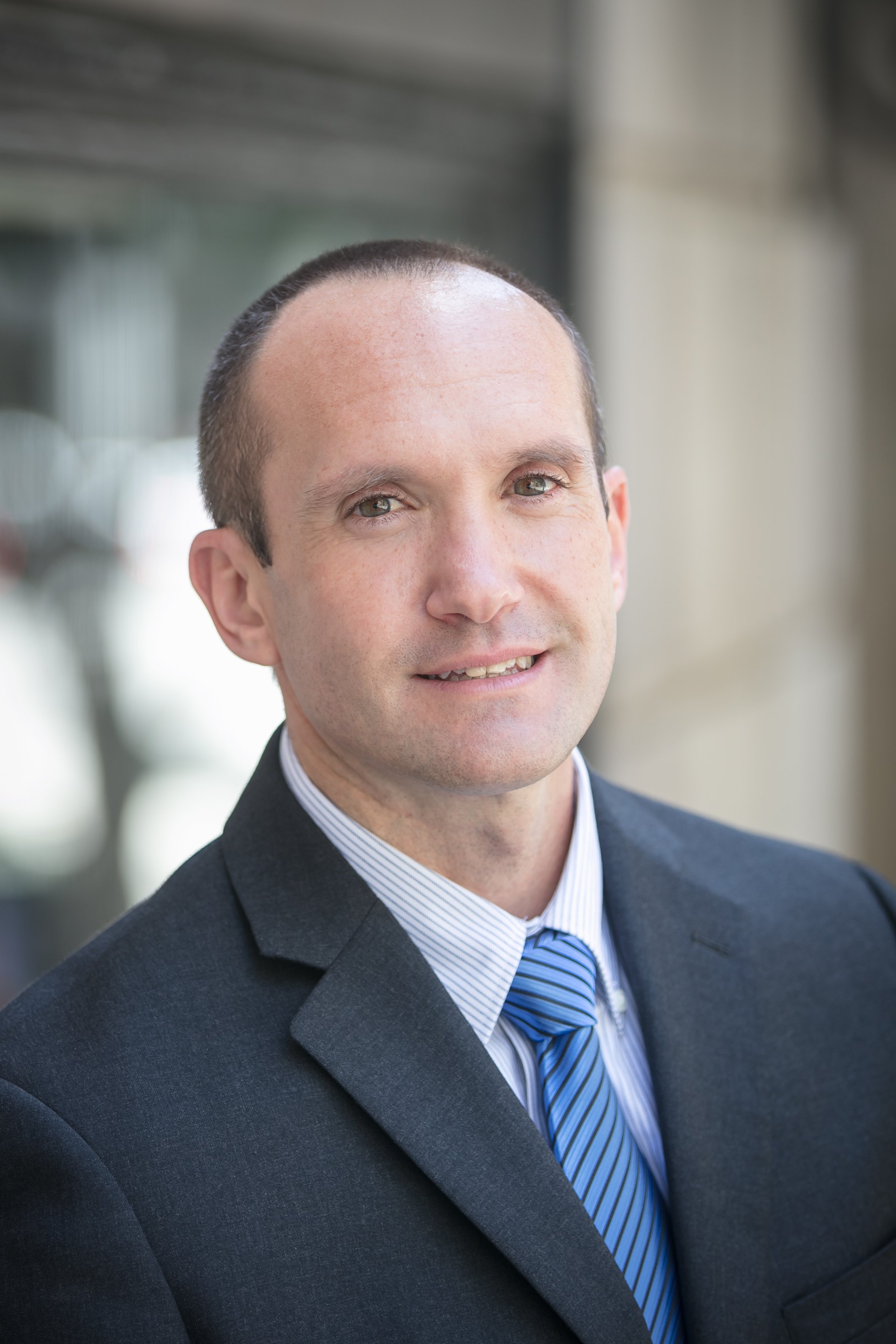 Portrait of a man in a dark suit with a light blue striped tie.