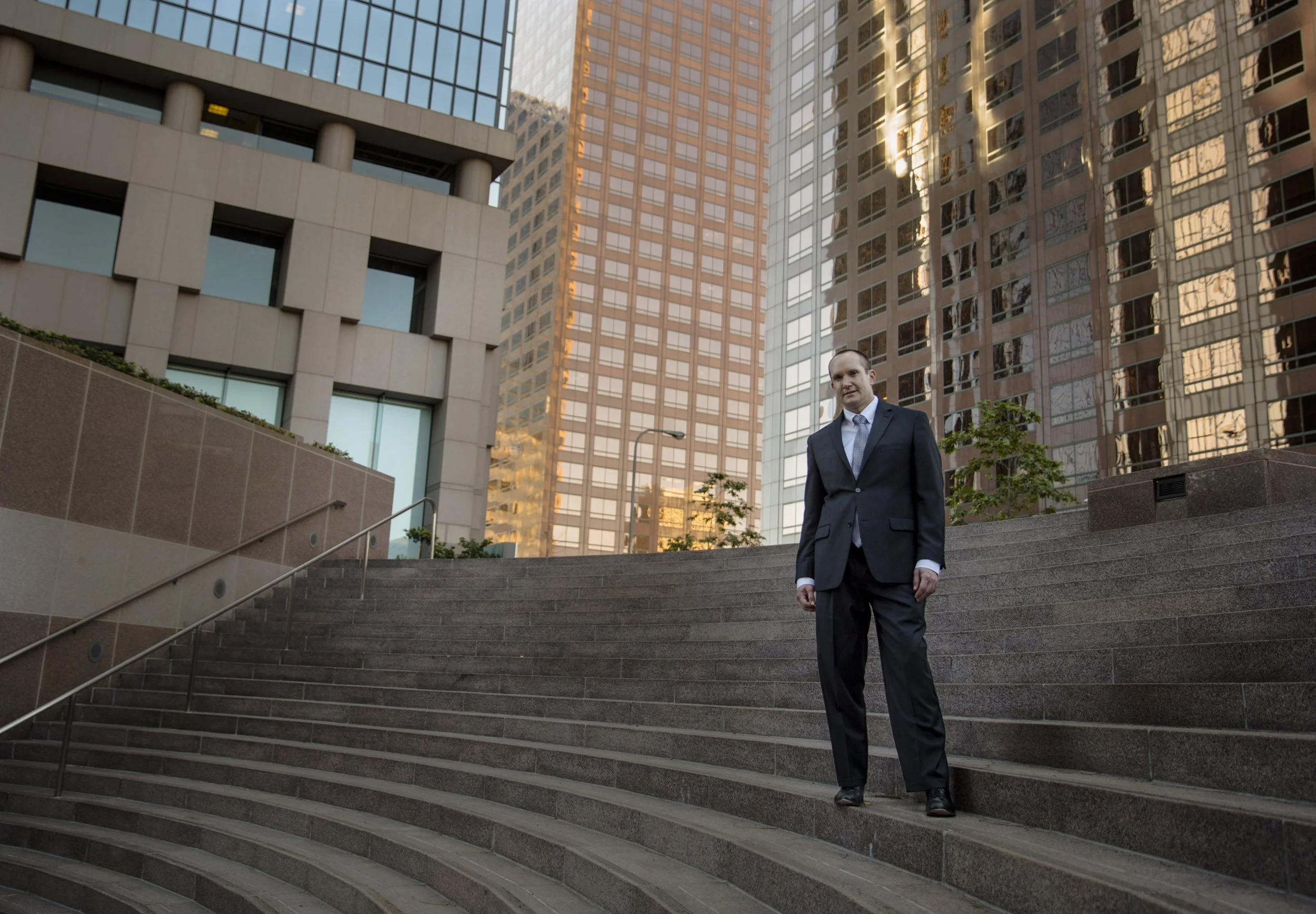 A man in a dark suit and tie walking down a wide outdoor staircase surrounded by modern office buildings with reflective glass windows in an urban cityscape.