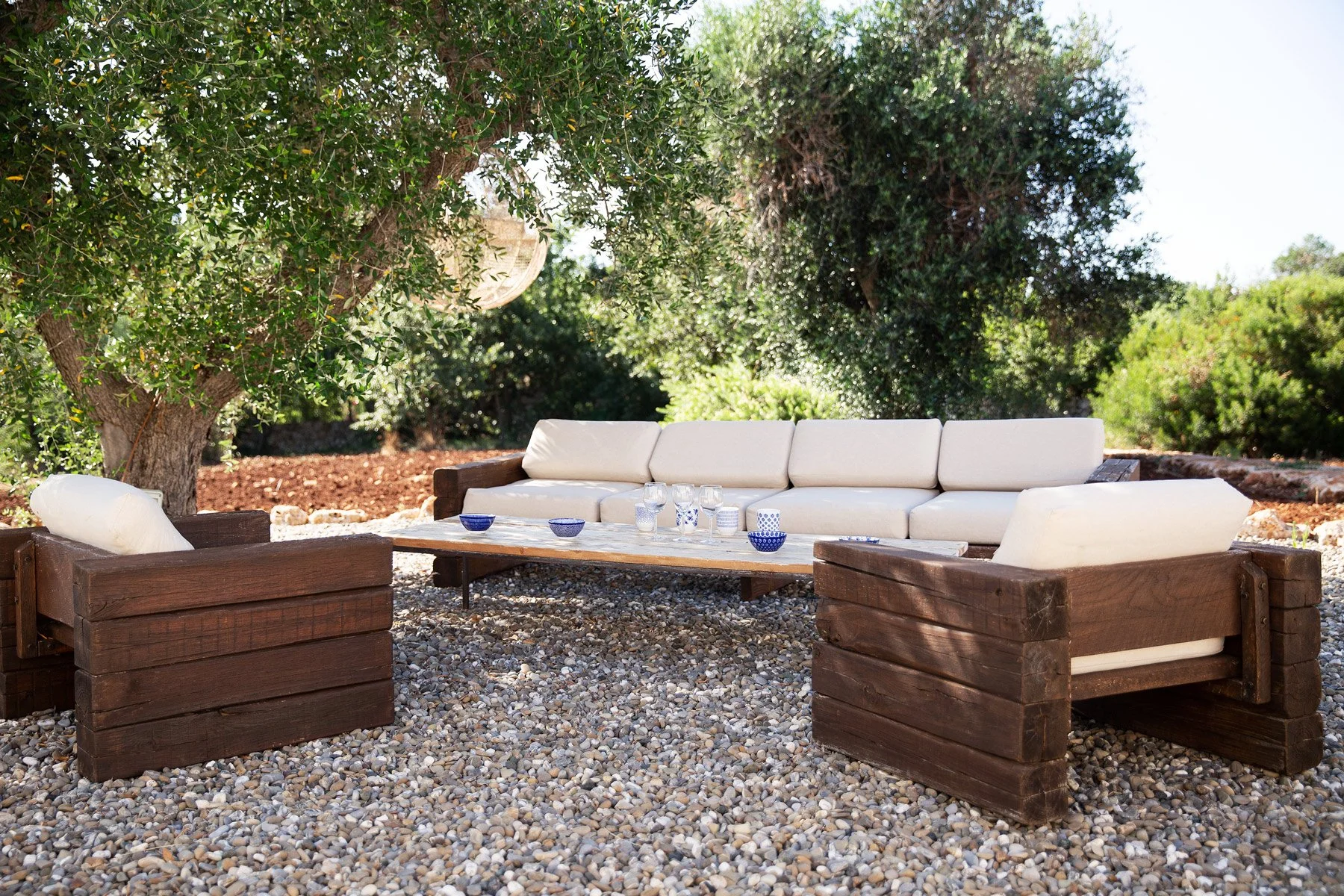 Un mobilier d'extérieur en bois avec des coussins blancs, placé sur un sol en galets, sous un arbre avec feuillage vert dense, avec des verres et des bols en céramique sur la table.