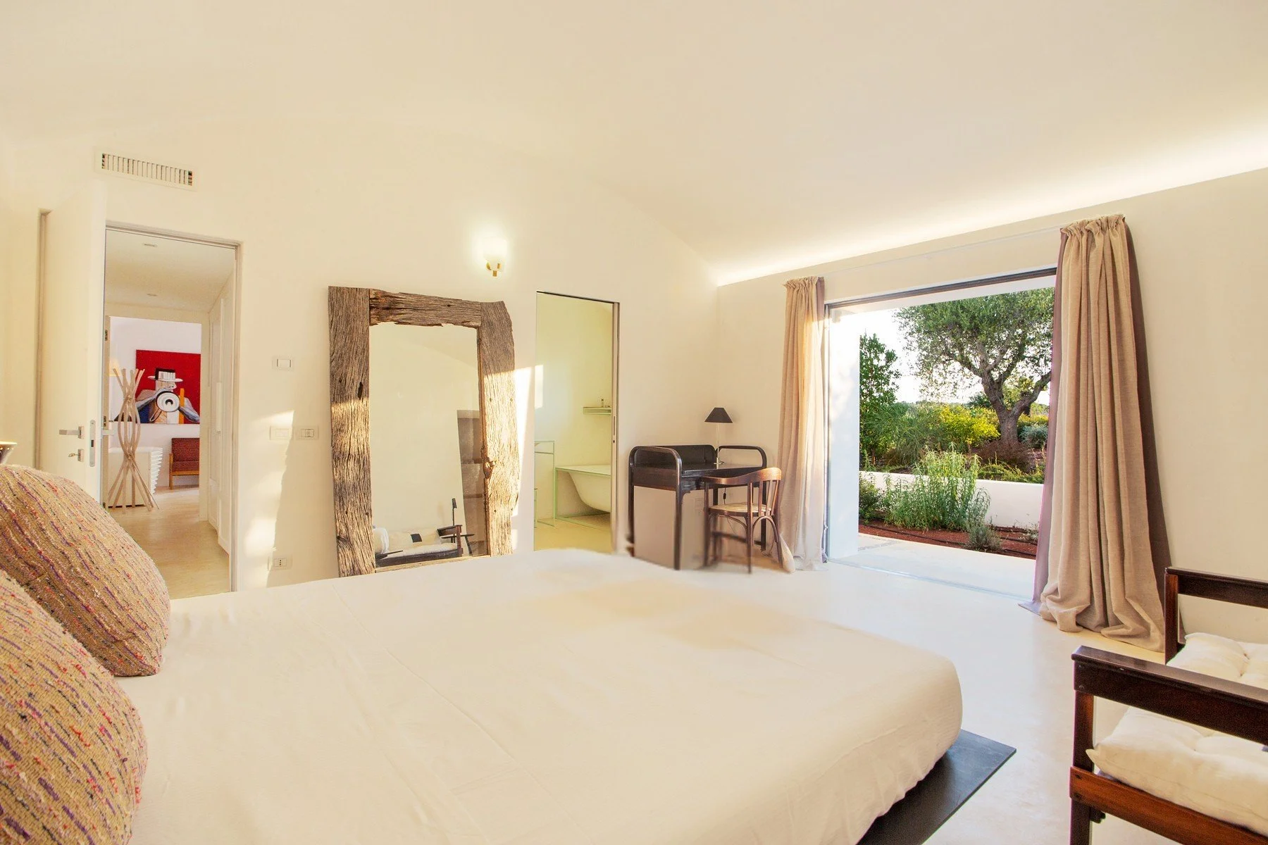 Chambre lumineuse avec un lit blanc, grande fenêtre donnant sur un jardin, porte ouverte vers une salle de bain, meubles en bois, lampes, et décor naturel.