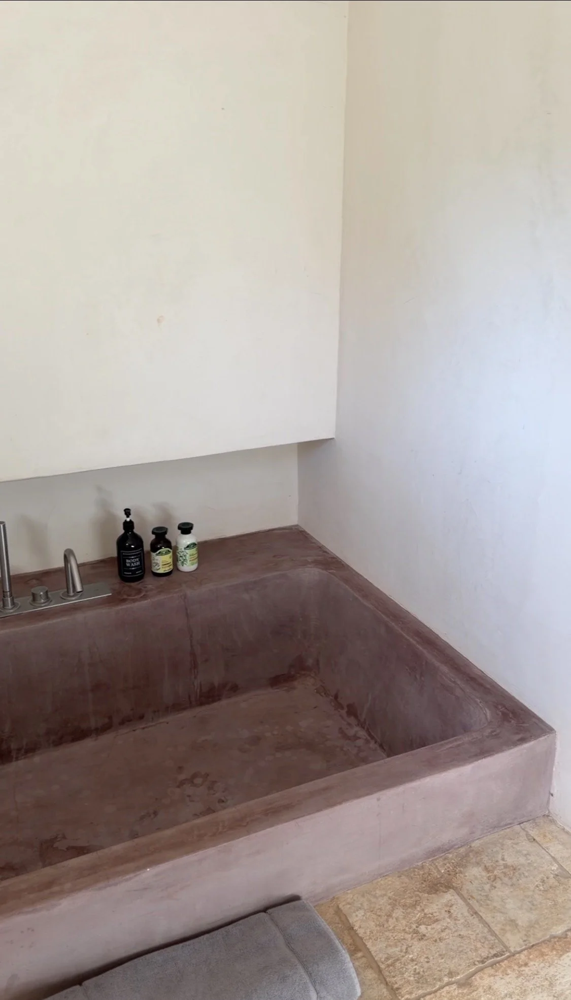 Baignoire en marbre marron avec trois bouteilles de savon ou lotion sur le rebord, murs blancs, sol en carreaux beige.