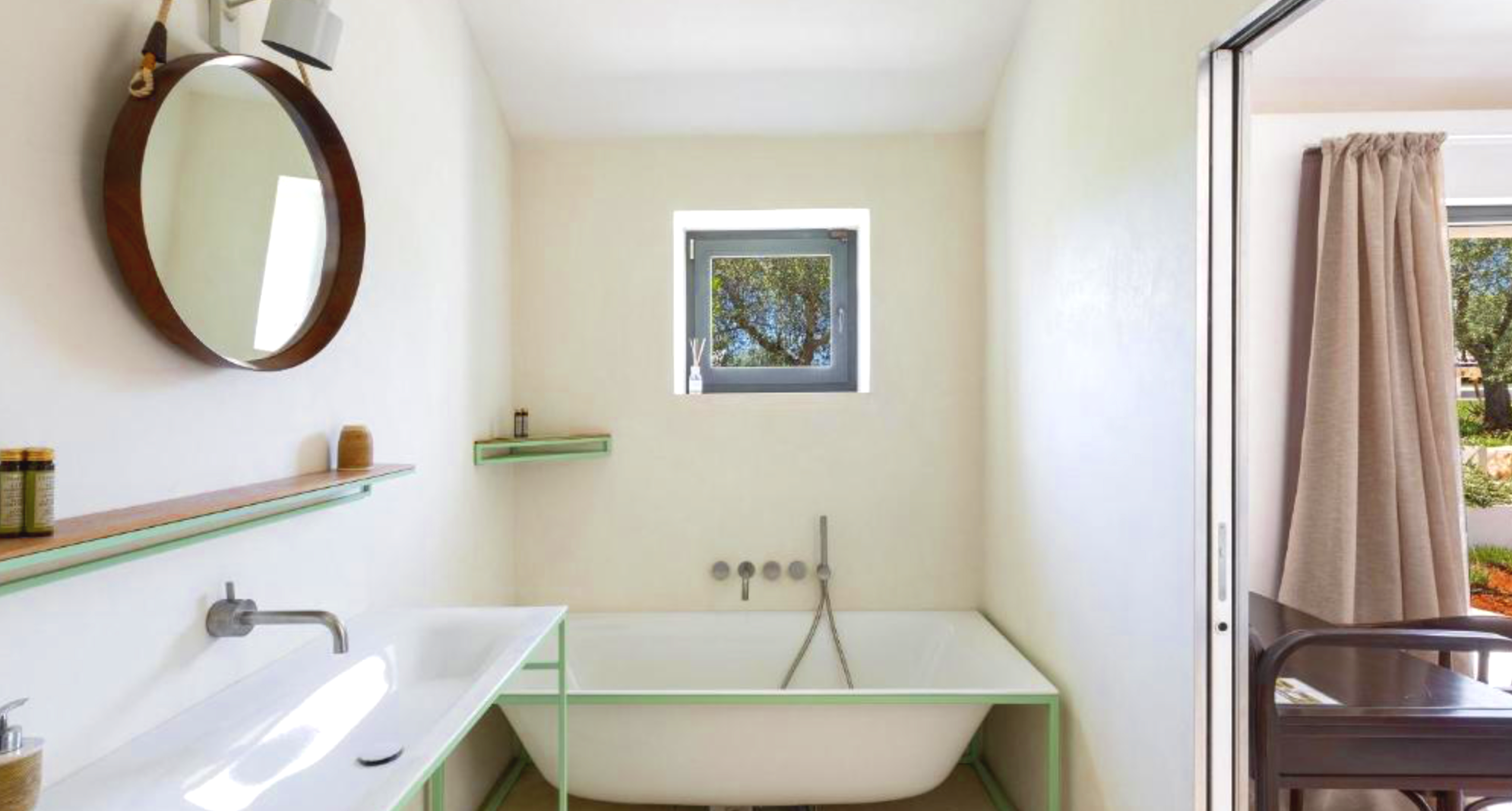 Une pièce de bain en cours de rénovation avec une baignoire ancienne blanche, un lavabo blanc avec un robinet en métal, une petite fenêtre laissant entrer la lumière naturelle, une étagère en verre verte, un miroir rond en bois accroché au mur, des m