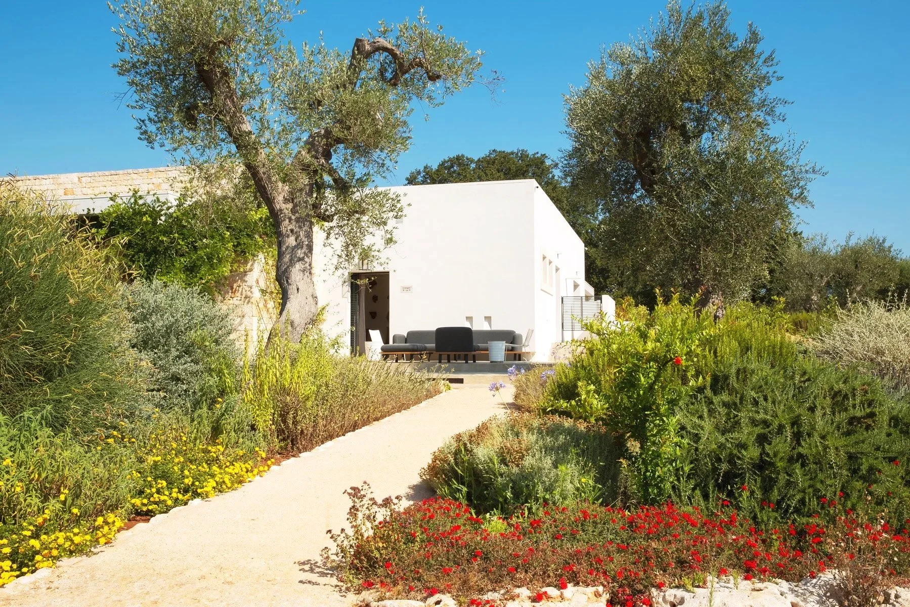 Une maison blanche moderne avec un mobilier extérieur, entourée de jardins fleuris et d'arbres, sous un ciel bleu clair.
