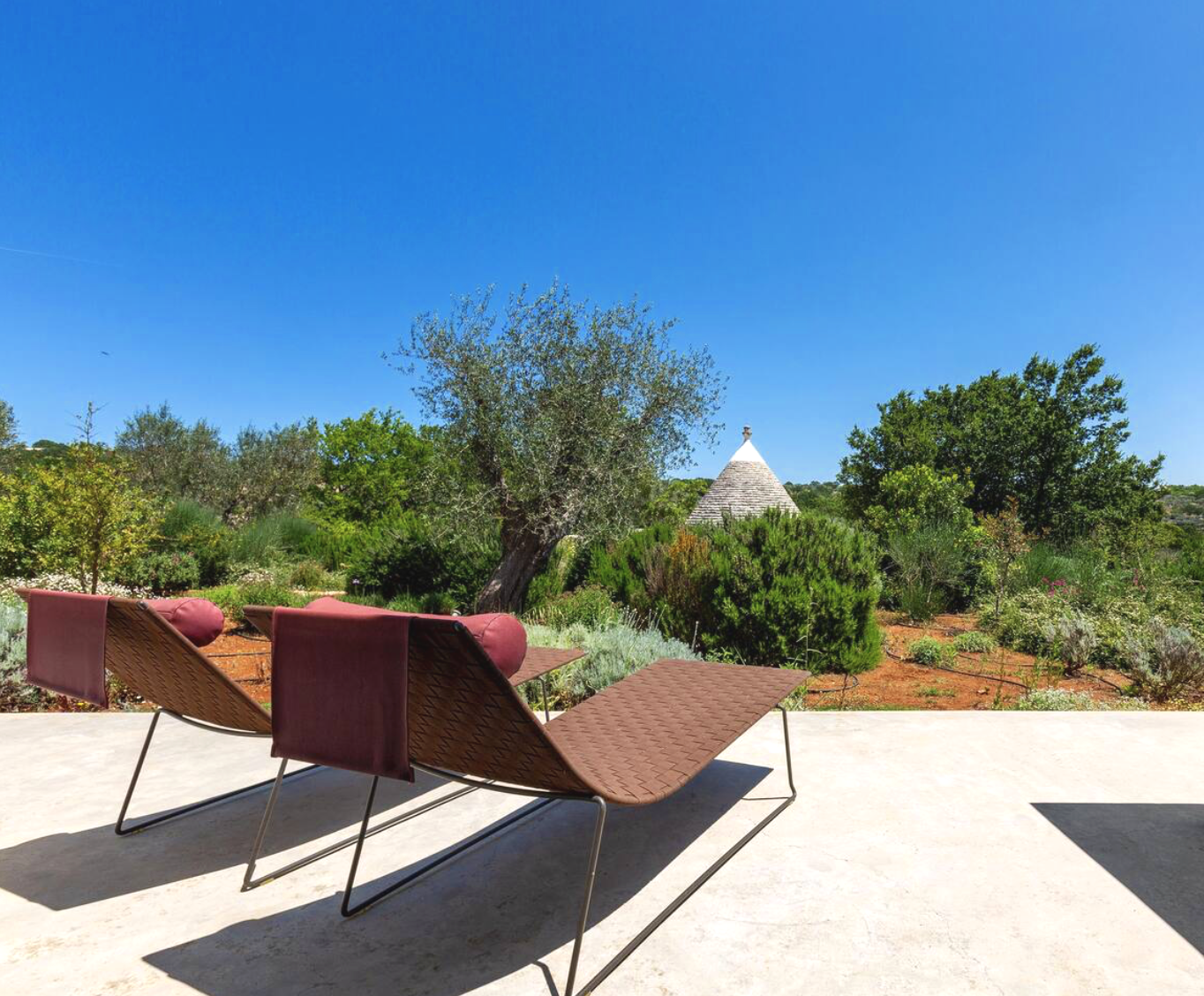 Chaises longues en bois et en métal sur une terrasse en pierre, vue sur un jardin avec des arbres et un bâtiment en forme de cône blanc en arrière-plan sous un ciel bleu clair.
