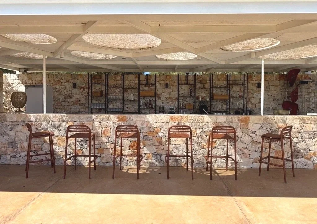 Un bar en extérieur avec une façade en pierre, six tabourets en bois, un plafond blanc avec des décorations rondes, et un mur en pierre avec des étagères en métal et des objets divers.