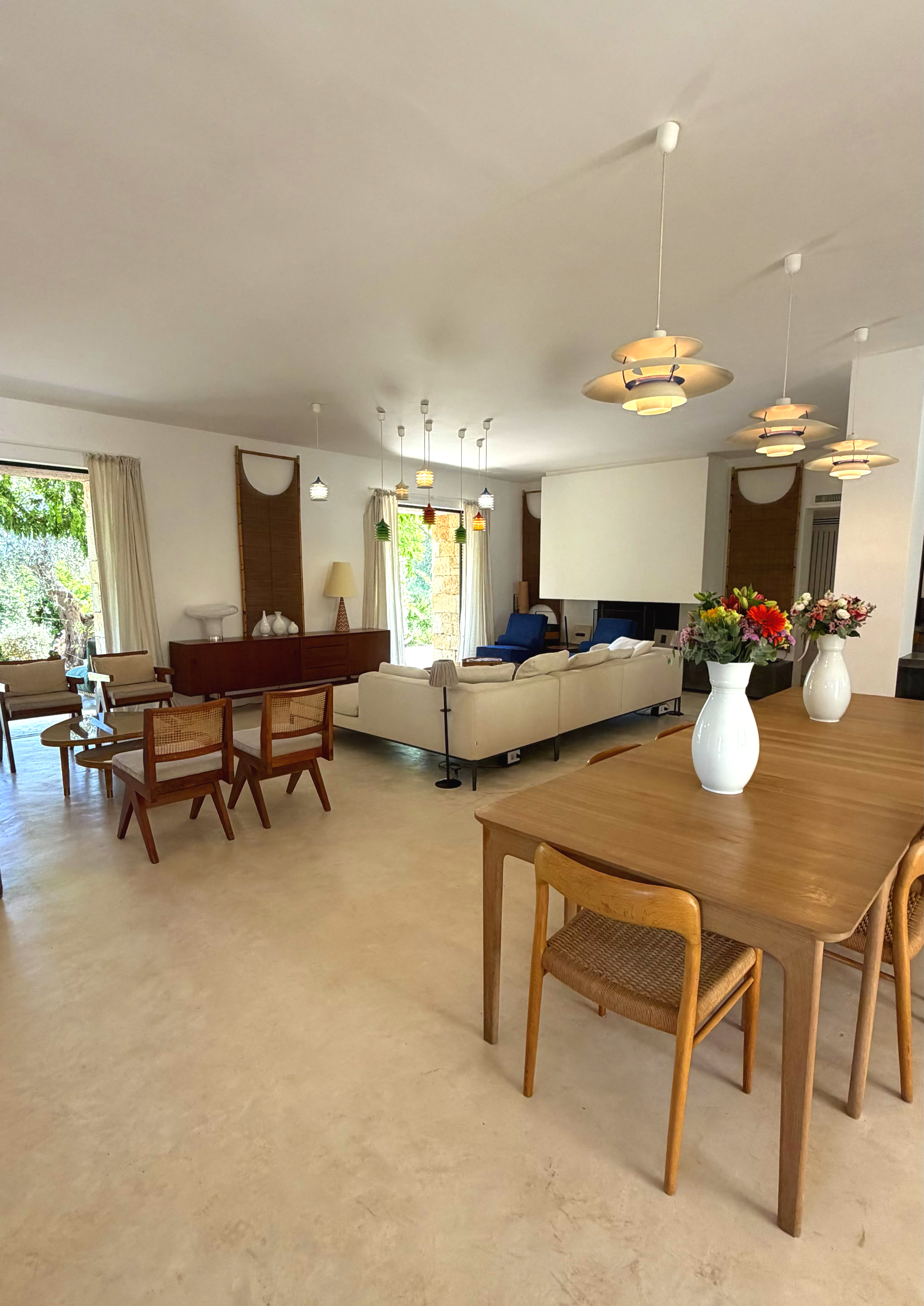 Salon spacieux avec table en bois, chaises en bois avec assise en cannage, canapés blancs, lampes suspendues modernes, vases avec fleurs colorées, grande fenêtre avec rideaux, décor contemporain.