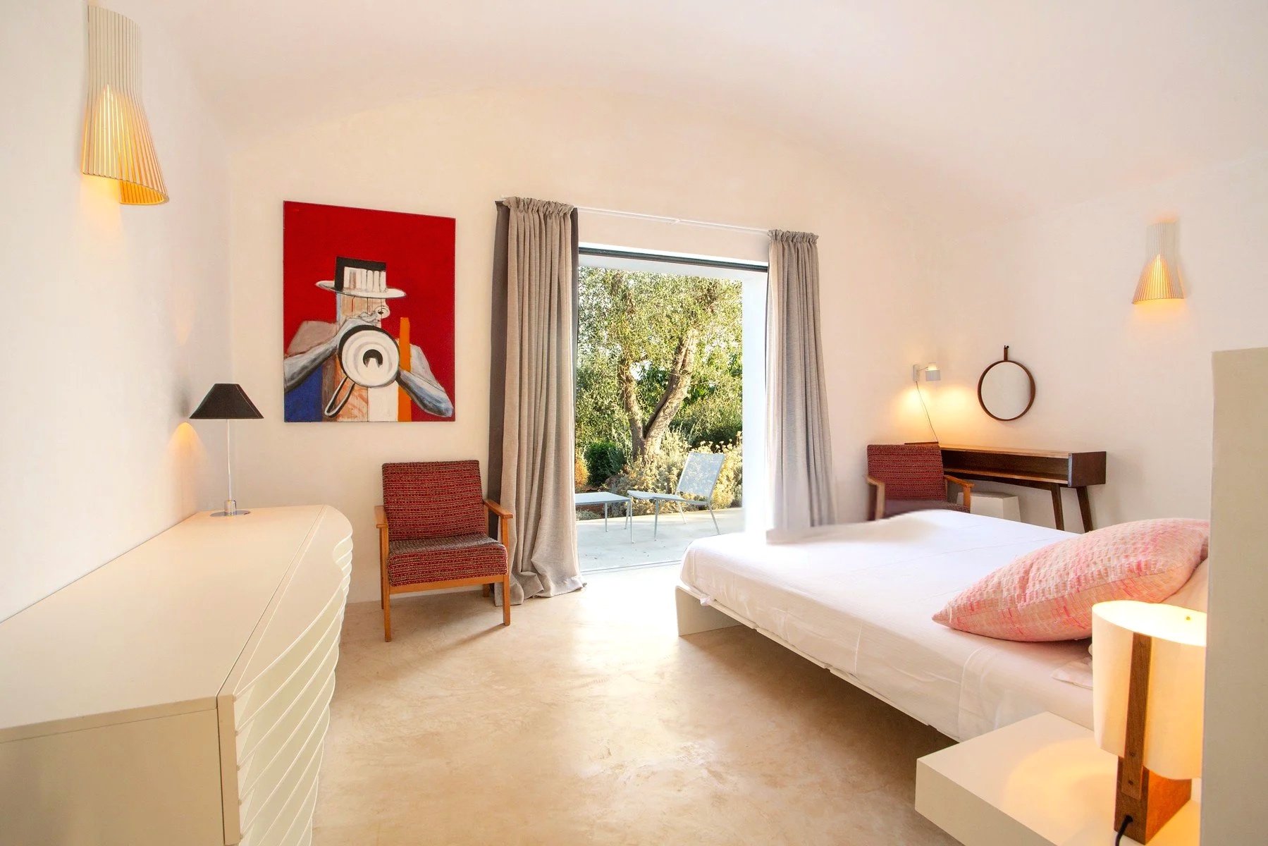 Chambre d'hôtel lumineuse avec un lit, deux chaises, une porte-fenêtre donnant sur un patio avec un arbre. La chambre est décorée avec une peinture abstraite, des lampes murales, un miroir rond et une petite table avec un lampadaire.