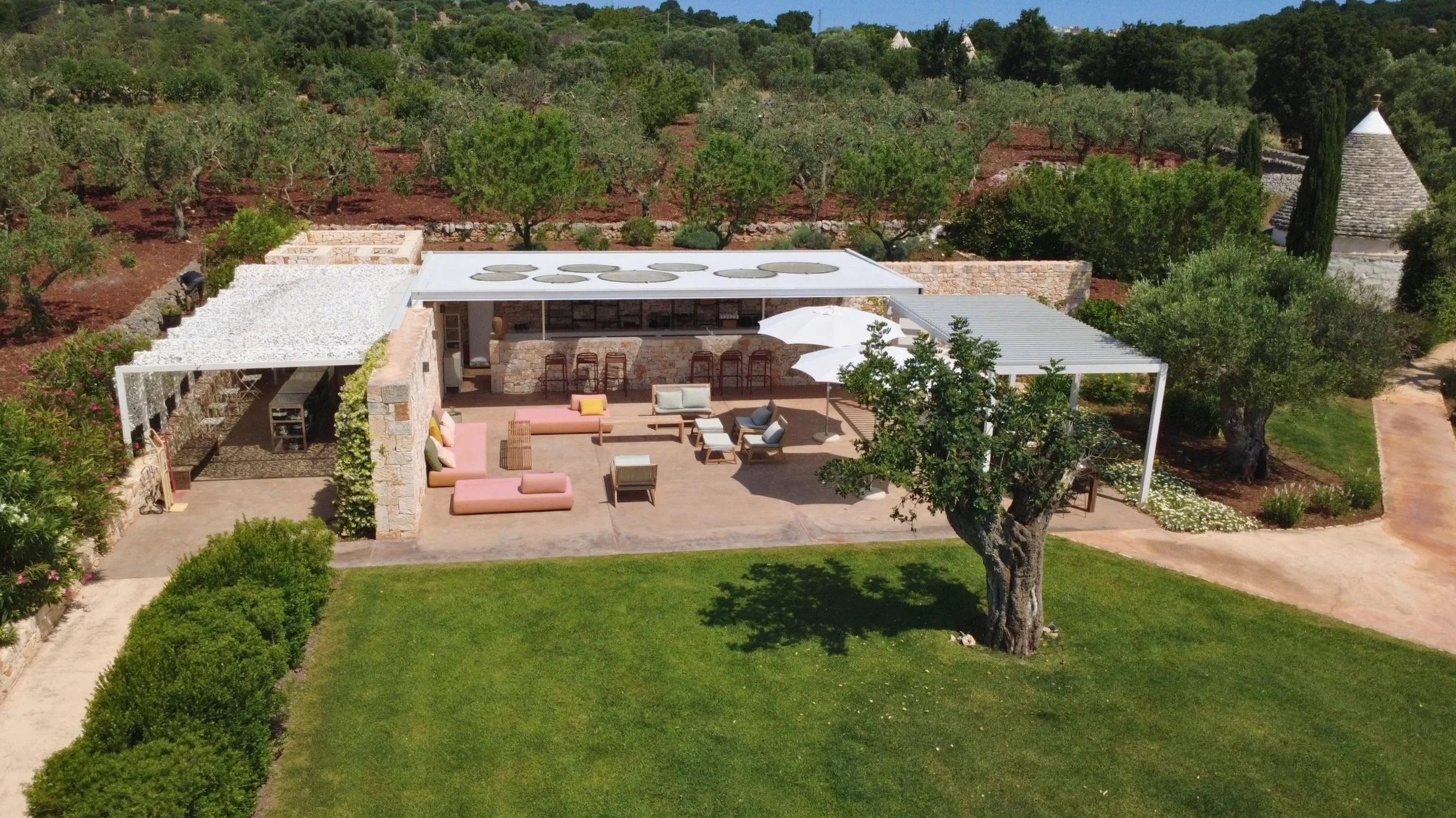 Une maison moderne avec une terrasse extérieure équipée de canapés, tables et parasols, entourée de verdure et d'arbres, dans un paysage naturel avec des collines et des arbres en arrière-plan.