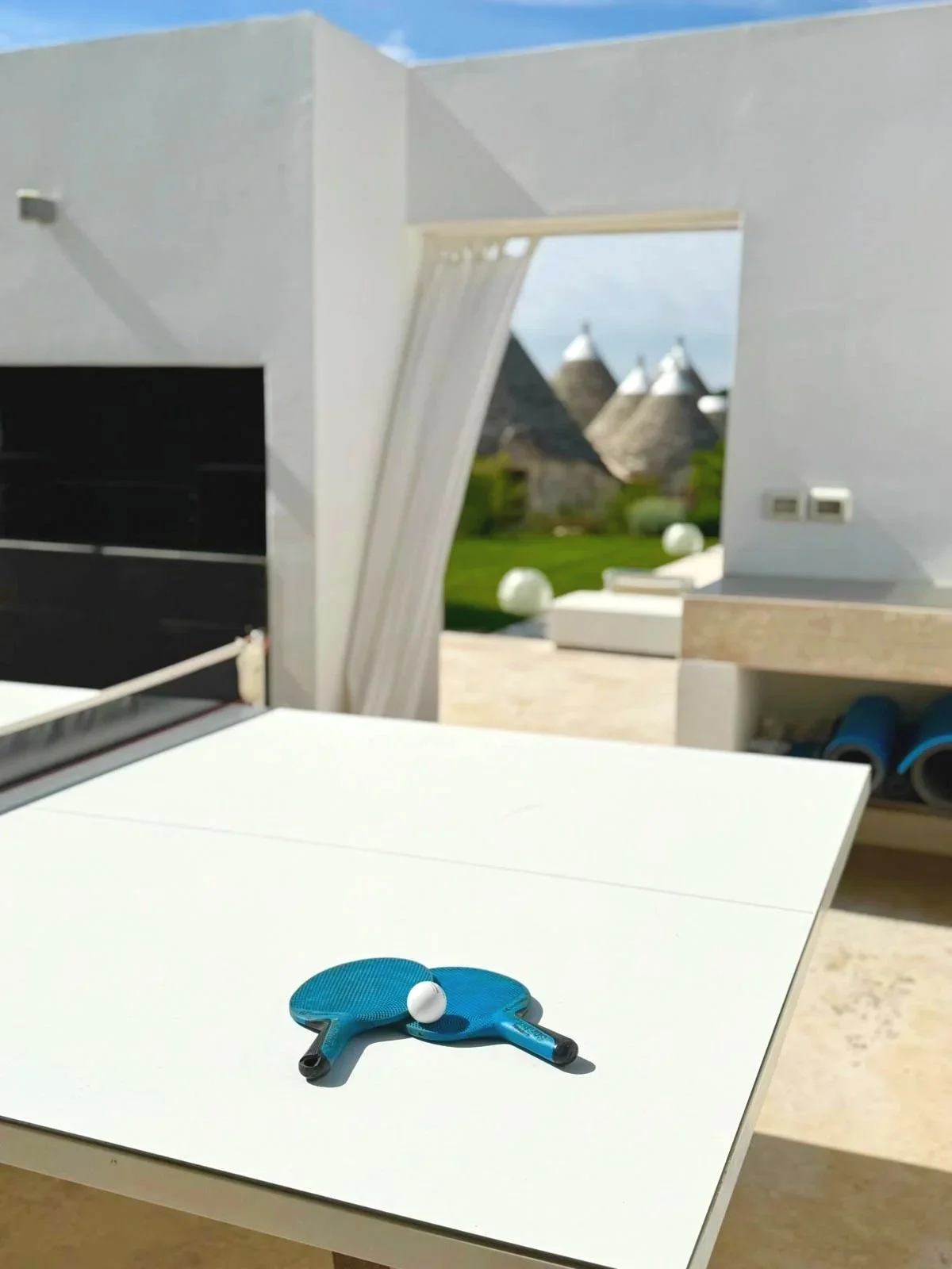 Une table de ping-pong avec une raquette bleue et une balle, située à l'extérieur sur une terrasse ensoleillée, avec des maisons de toit conique en pierre en arrière-plan.