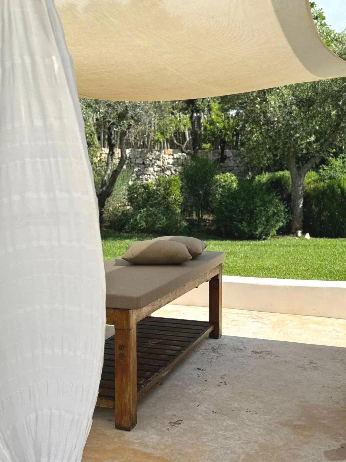 Une banquette en bois avec deux coussins beige à l'extérieur, près d'une surface en béton, avec un jardin et des arbres en arrière-plan, sous une grande toile beige.