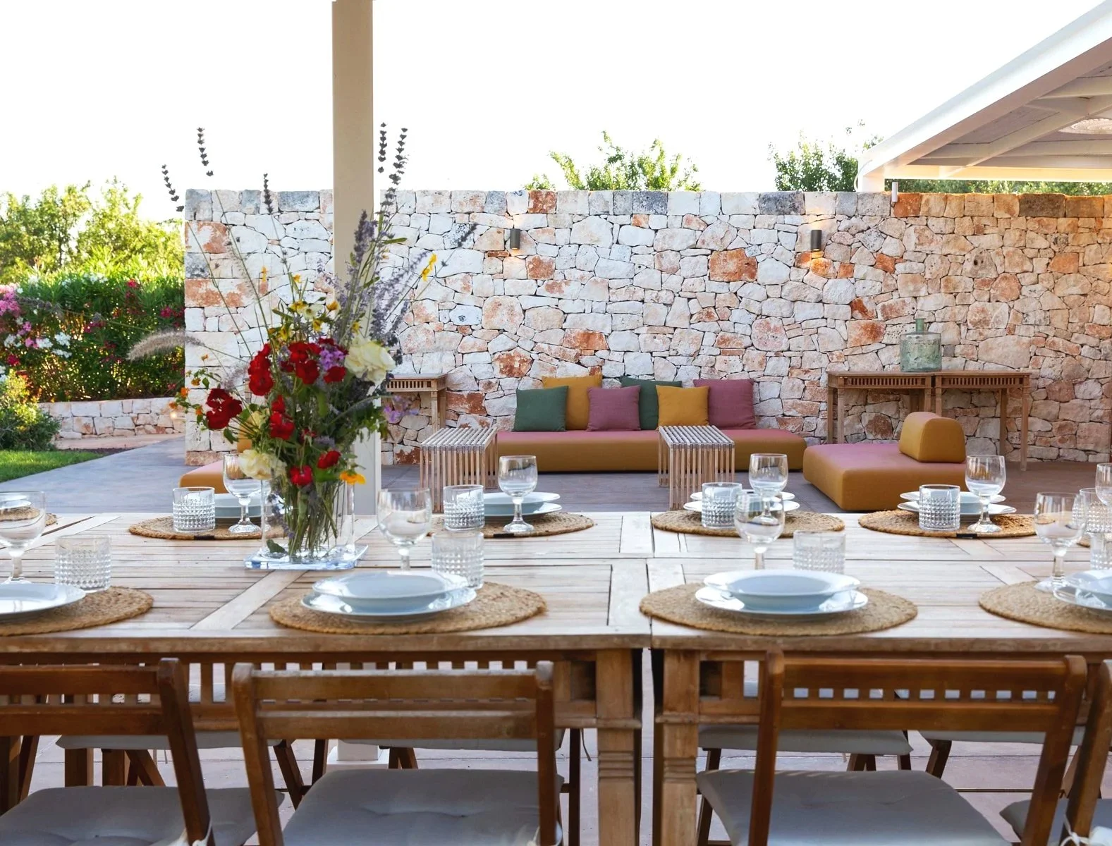 Une salle à manger extérieure avec une table en bois, des assiettes, des verres et des bougies. À l’arrière, un espace lounge avec un canapé pourvu de coussins colorés, une table en bois et un mur en pierre décoratif. Un vase avec des fleurs fraîches
